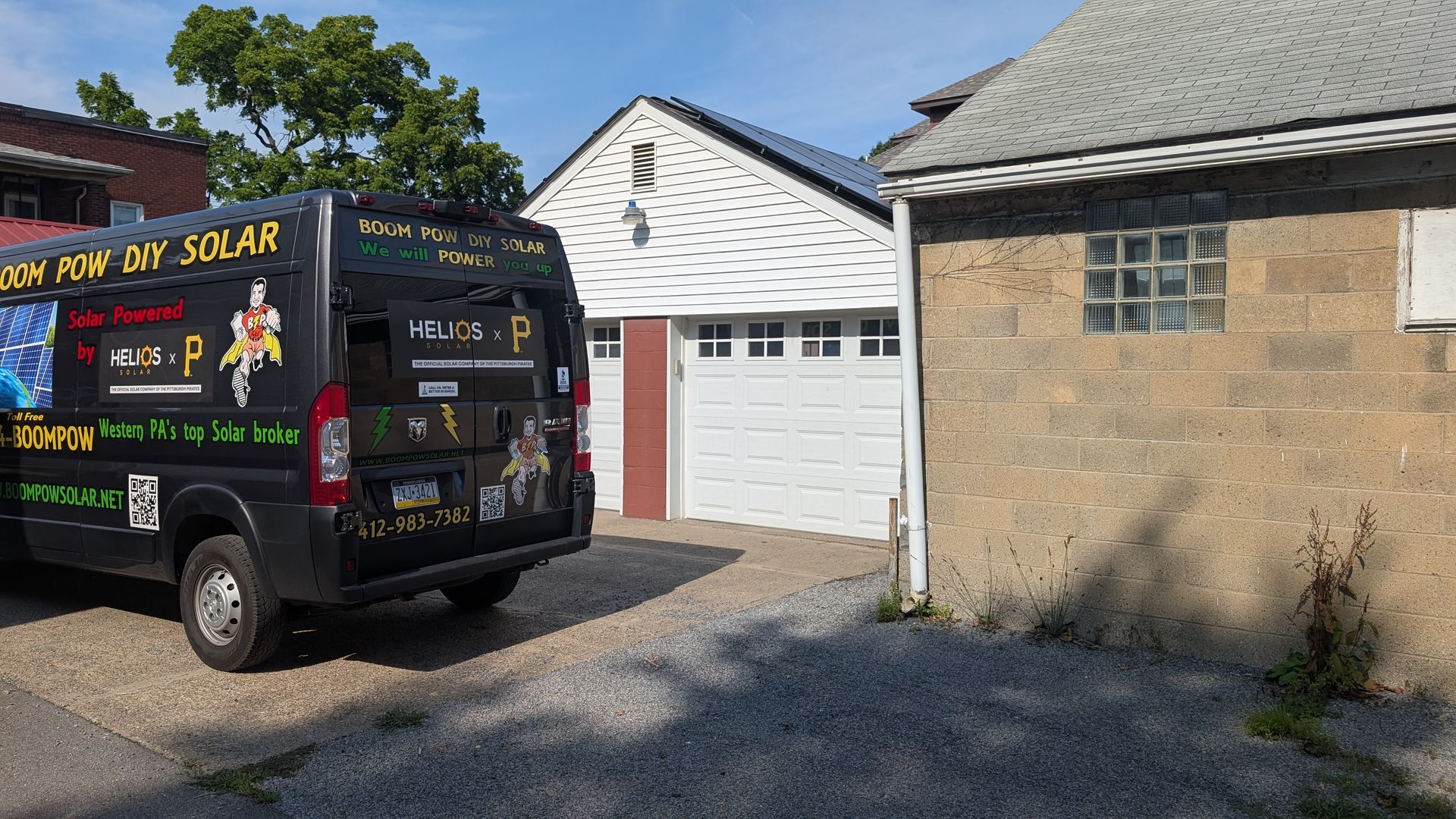 A black van with solar panel advertisements parked in front of a white garage and a gray cinder block building on a sunny day.