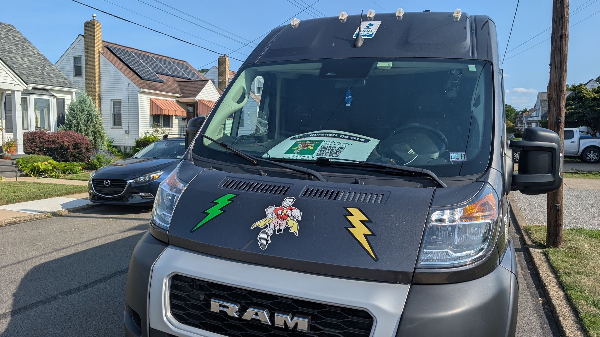 A dark gray RAM van with stickers on the hood parked on a residential street. Houses and a car are in the background.