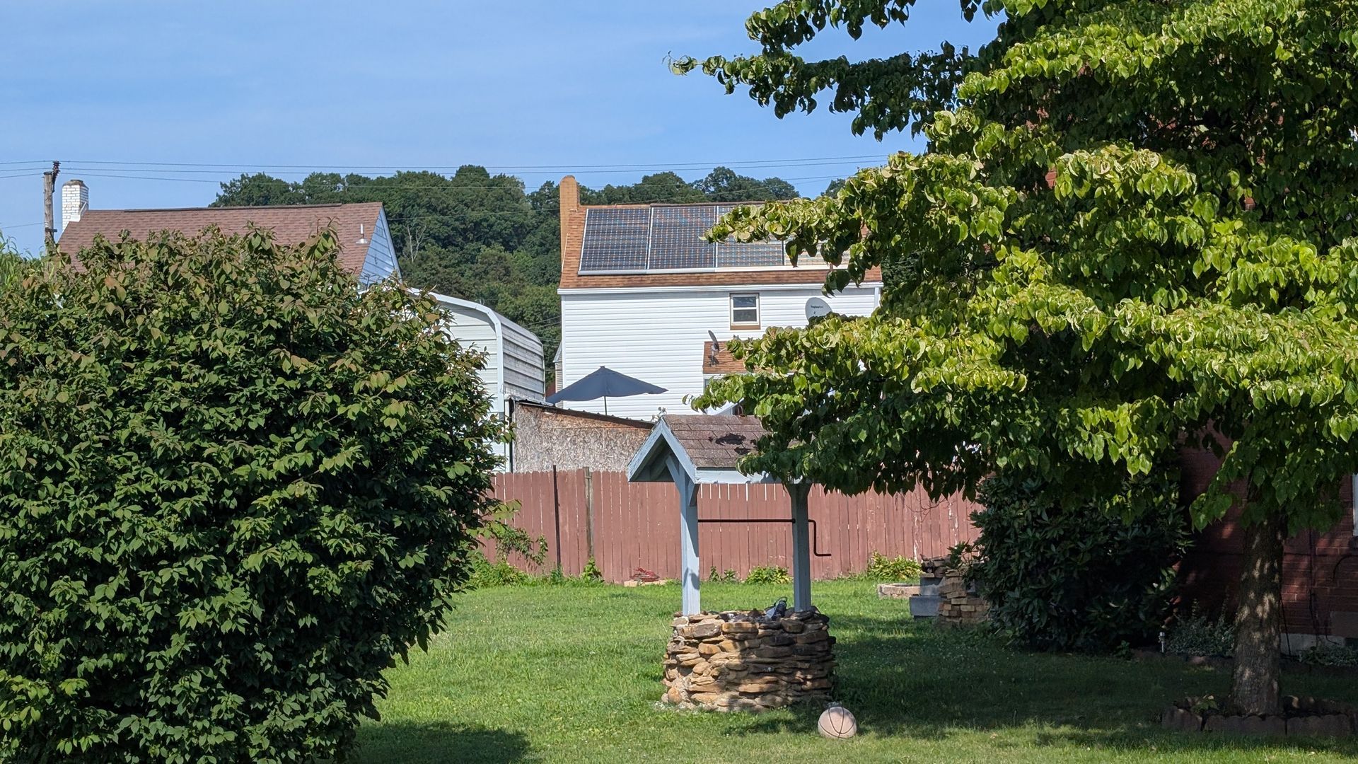 Lawn with a decorative well, green bushes, a weathered wooden fence, and a white building with a brown roof in the background.