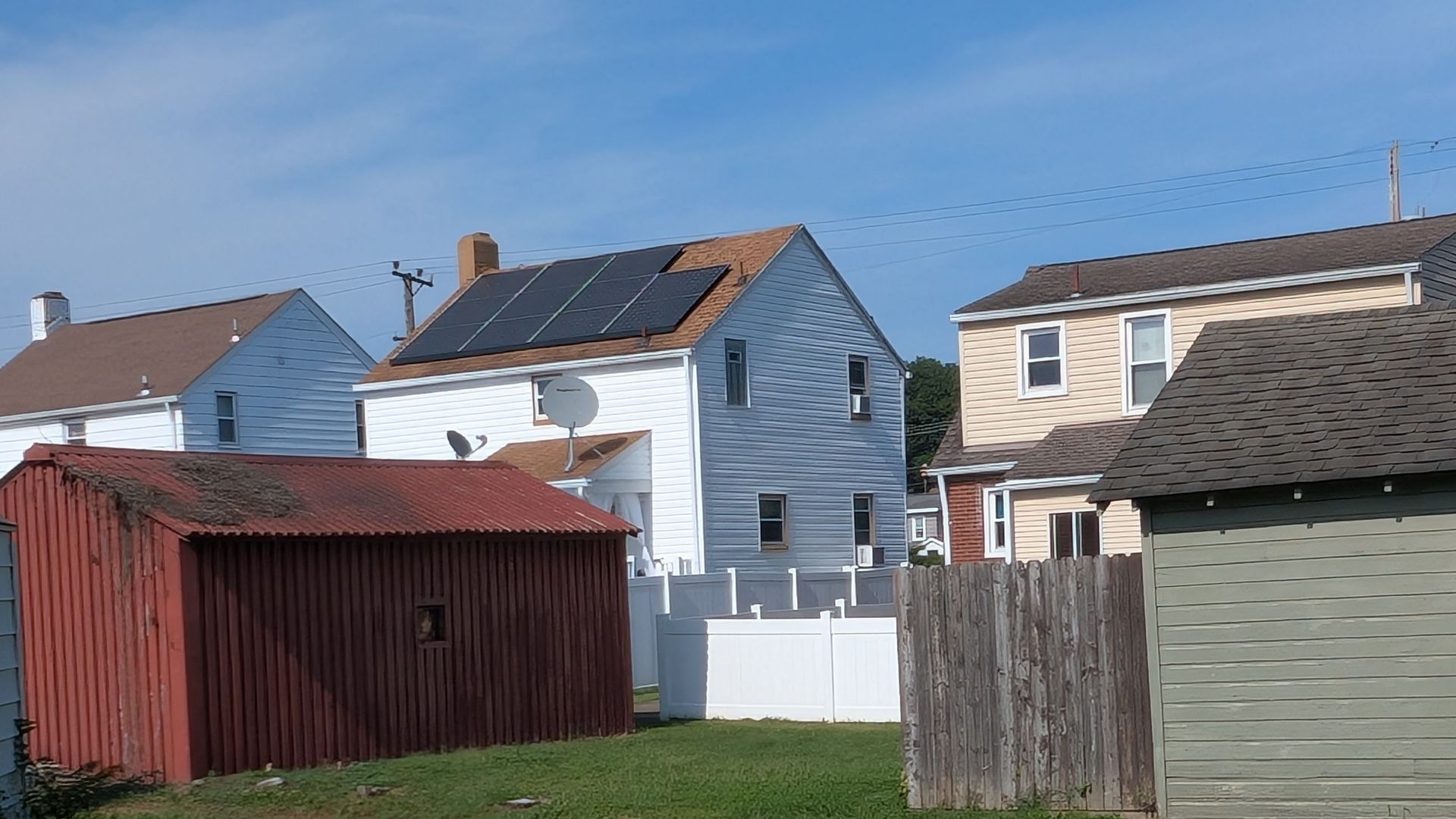 Backyards of houses with solar panels on one roof, blue sky overhead. A red shed in the foreground.
