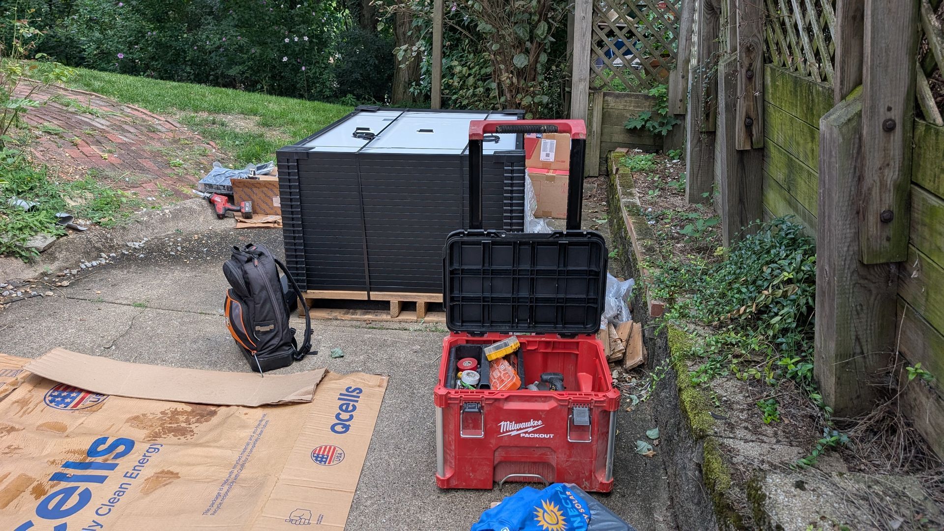 A red toolbox is open next to a pile of black panels and a backpack, with other equipment and cardboard, in a narrow outdoor area.