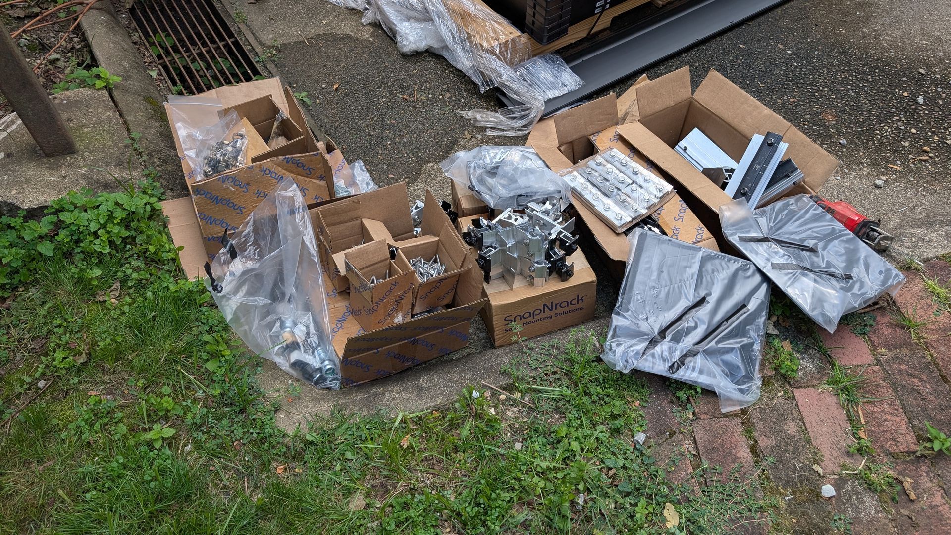 Cardboard boxes and wrapped items sit on grass and brick in an outdoor setting, likely containing components.