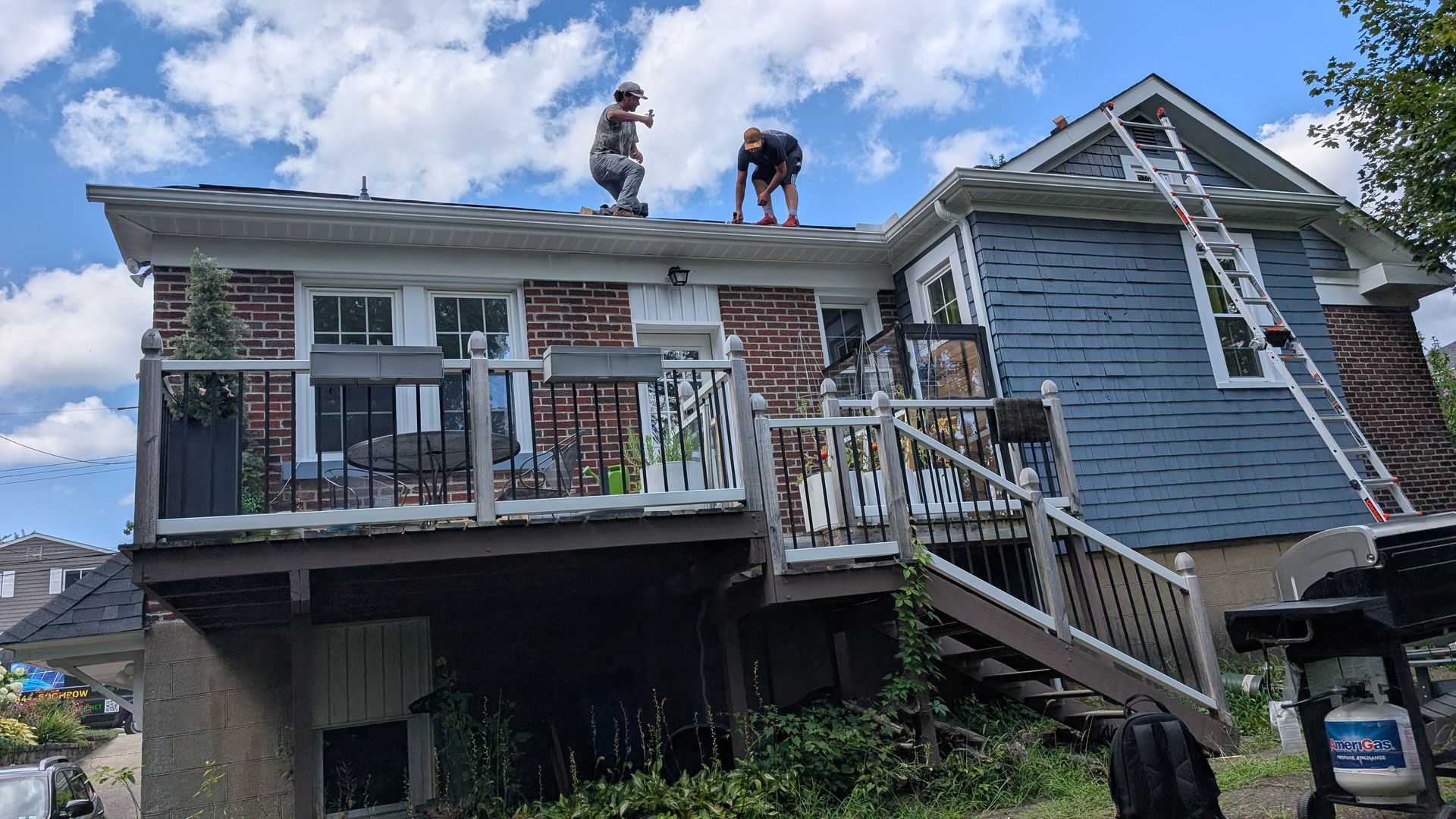 Two people working on a roof, one standing and one bending. House has blue siding and a deck with stairs. Ladder leaning against the side.