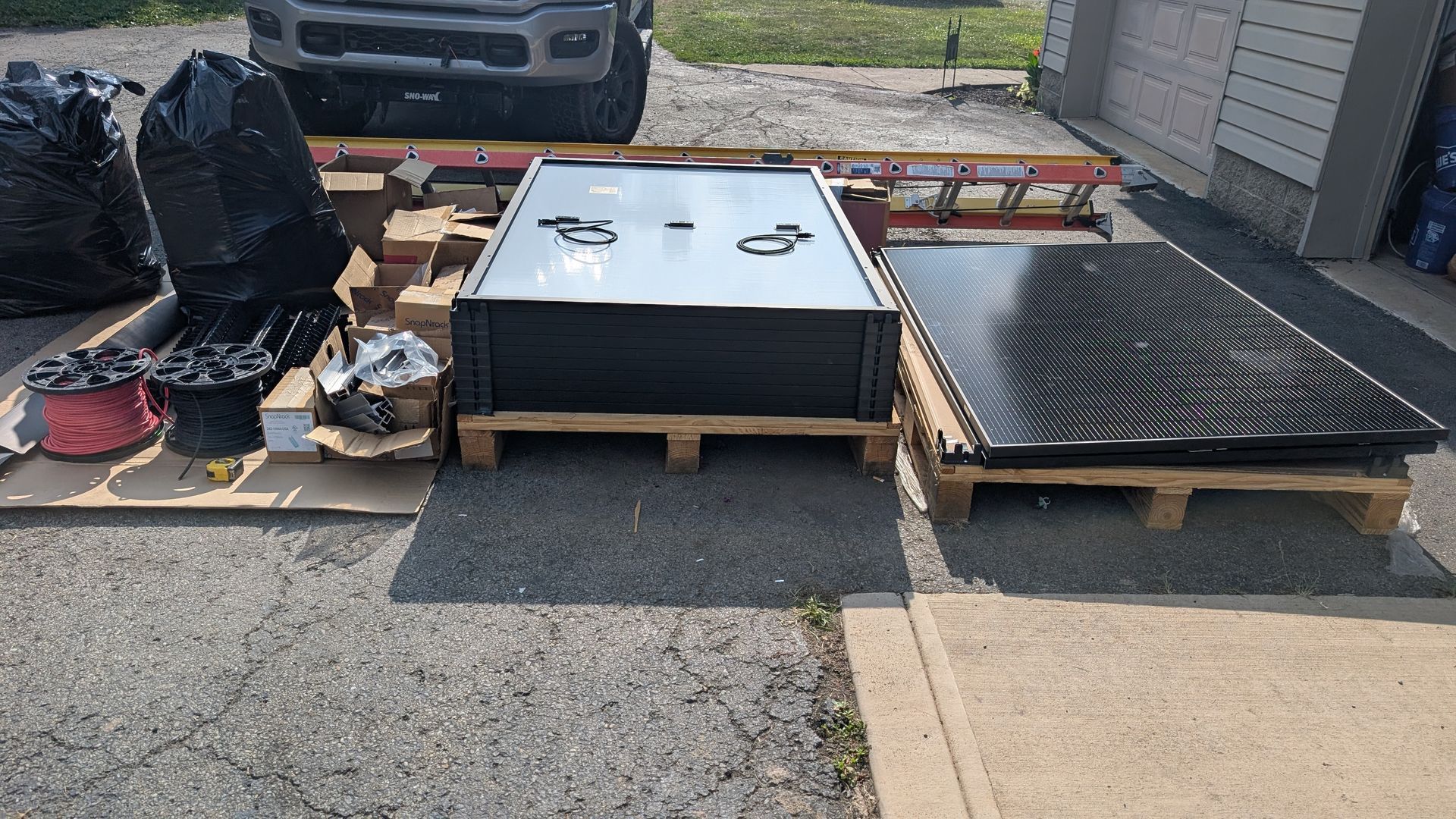 Solar panels stacked on pallets on a driveway, alongside a truck.