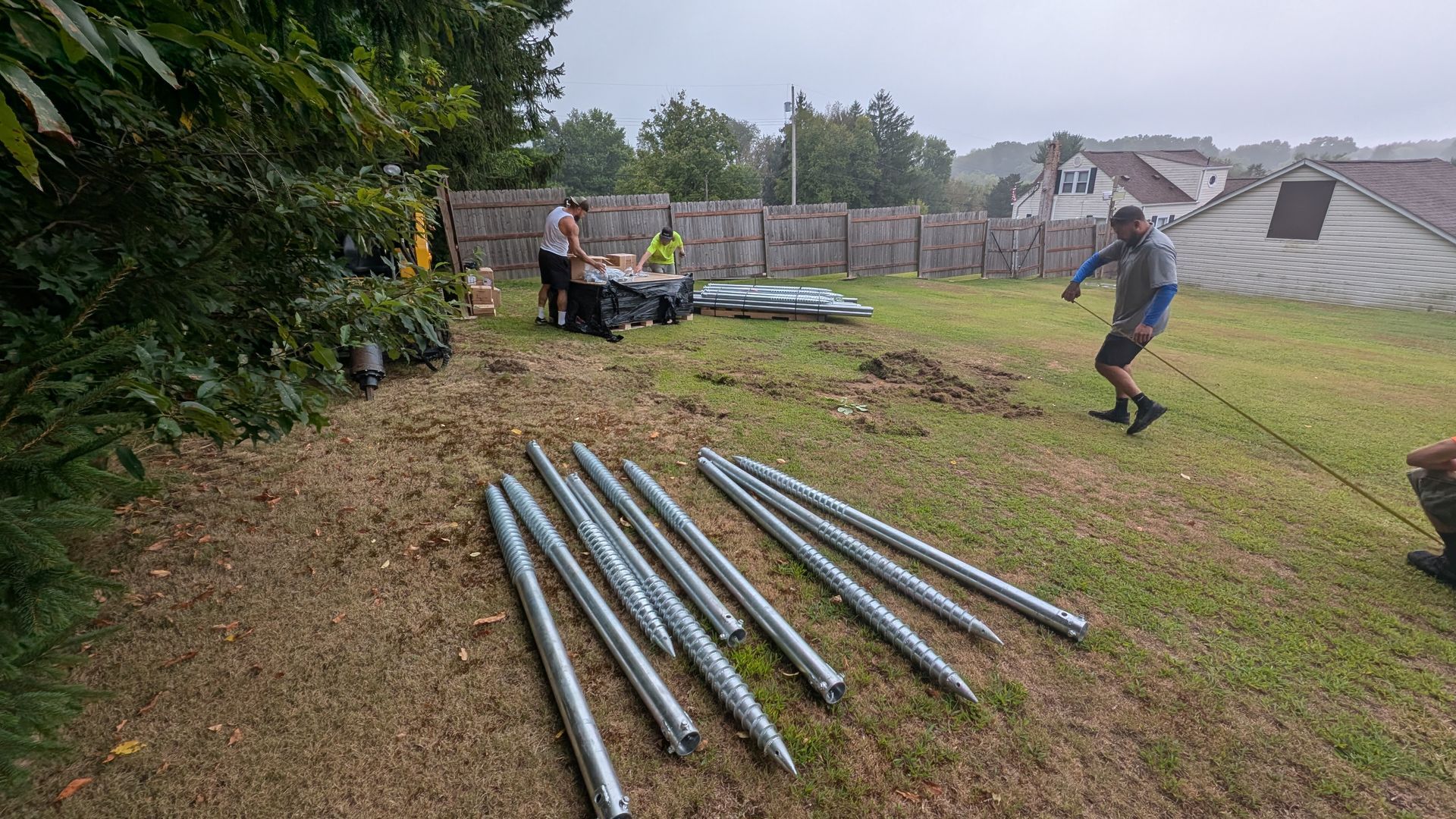 Workers installing tent supports on a grassy area, with tools and materials present, under overcast skies.