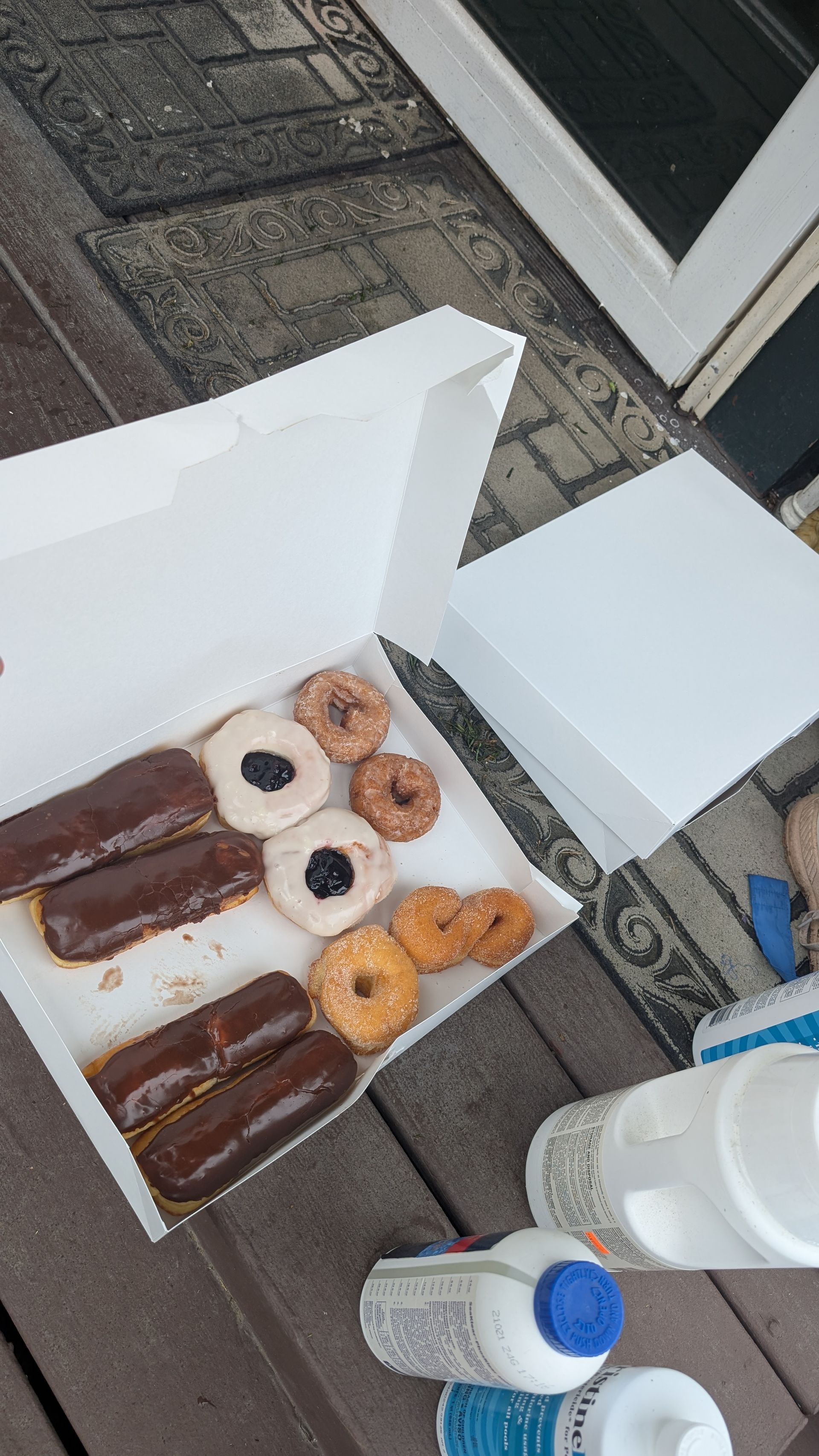 Box of donuts on a wooden deck; chocolate eclairs and assorted glazed donuts visible, near bottles of cleaning supplies.