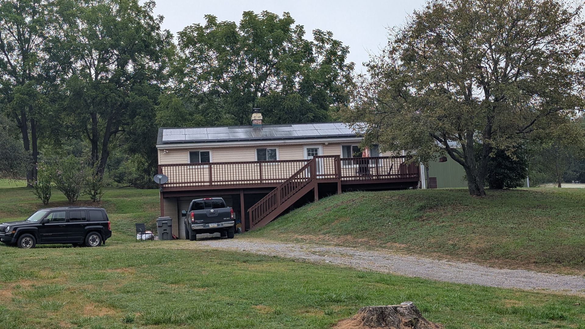 A house with a deck, two parked trucks, and trees on a grassy hill.