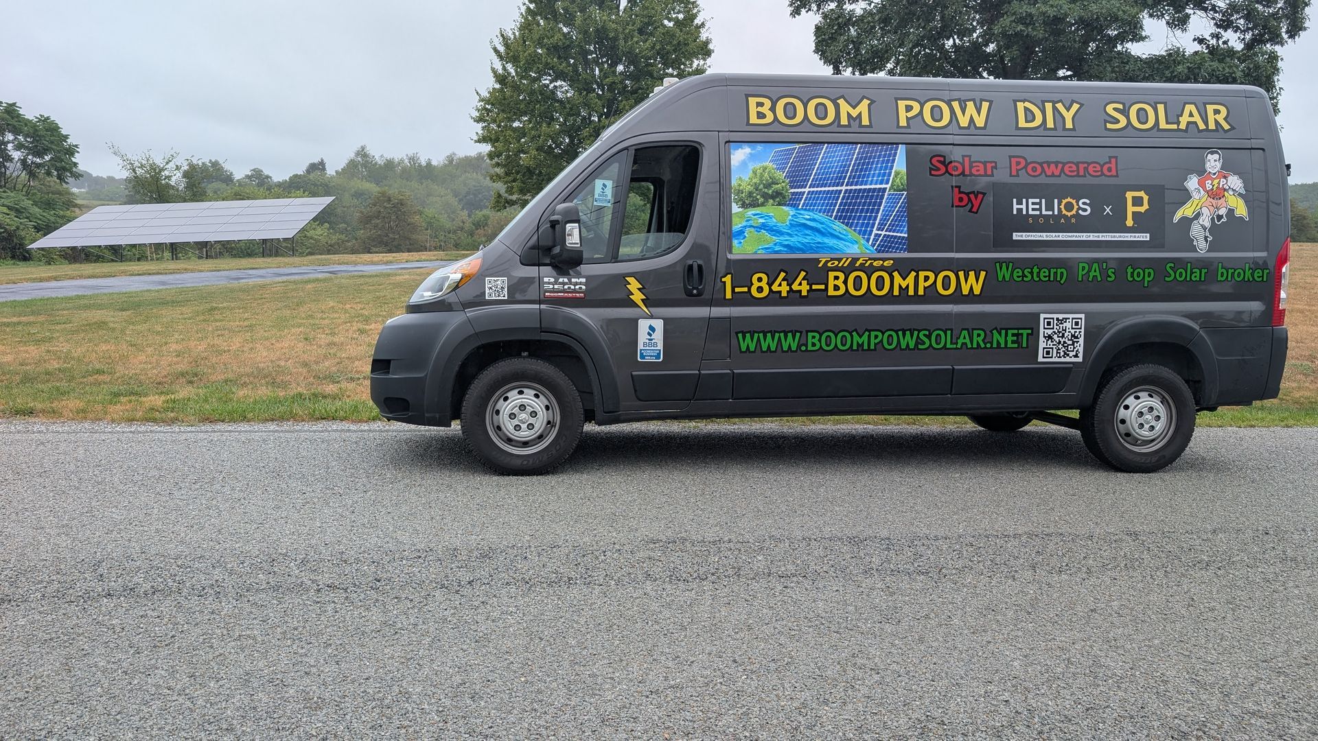 A dark van with Boom Wow DIY Solar signage parked next to solar panels on a gravel road.