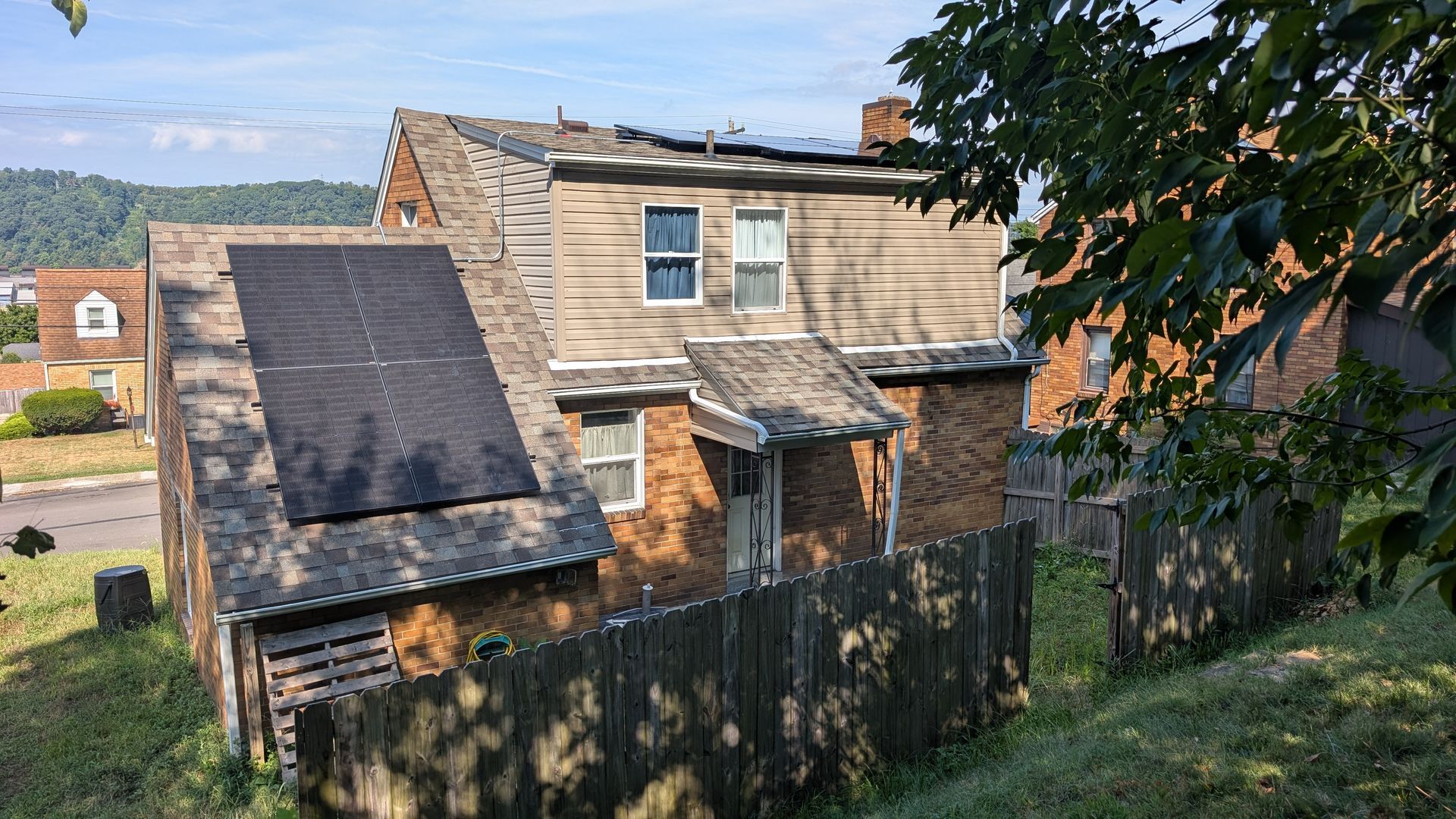 House with solar panels on roof, brick and wood siding, behind a wooden fence.