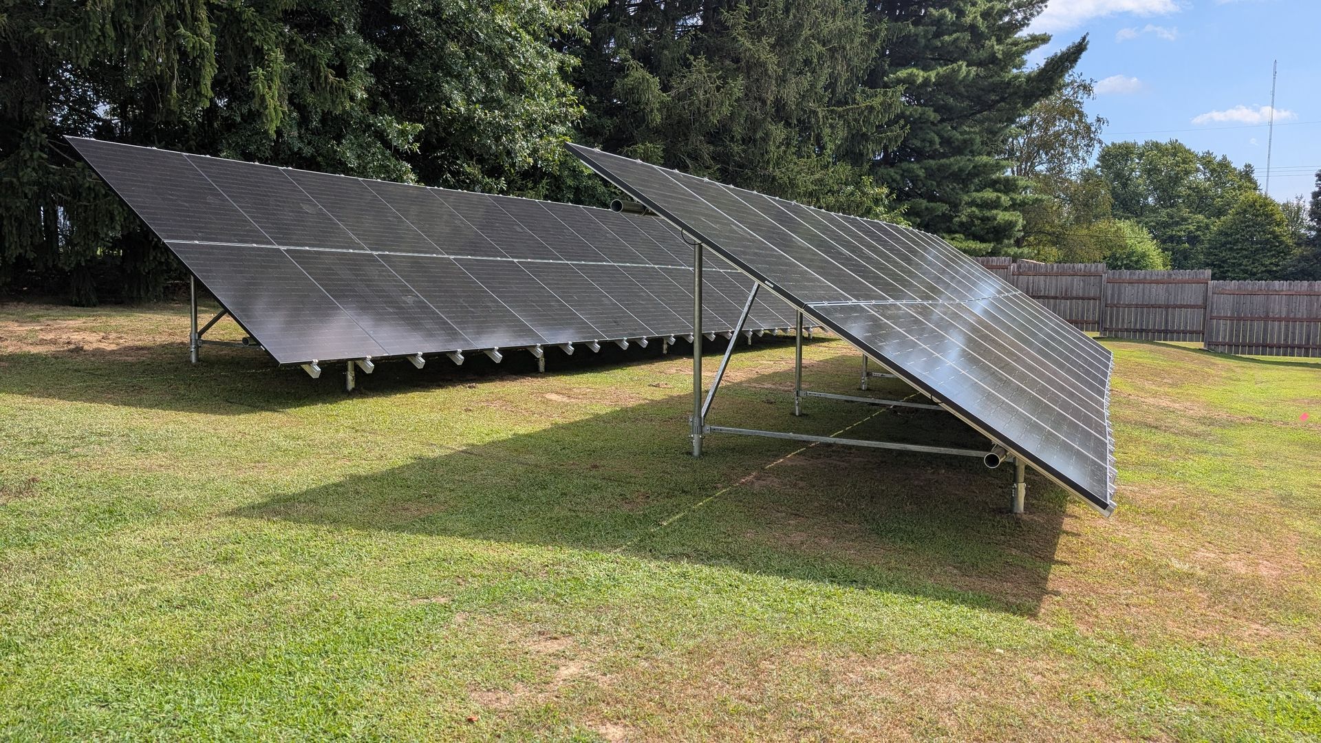 Solar panels arrayed in a grassy yard, angled towards the sun, with trees and a fence in the background.