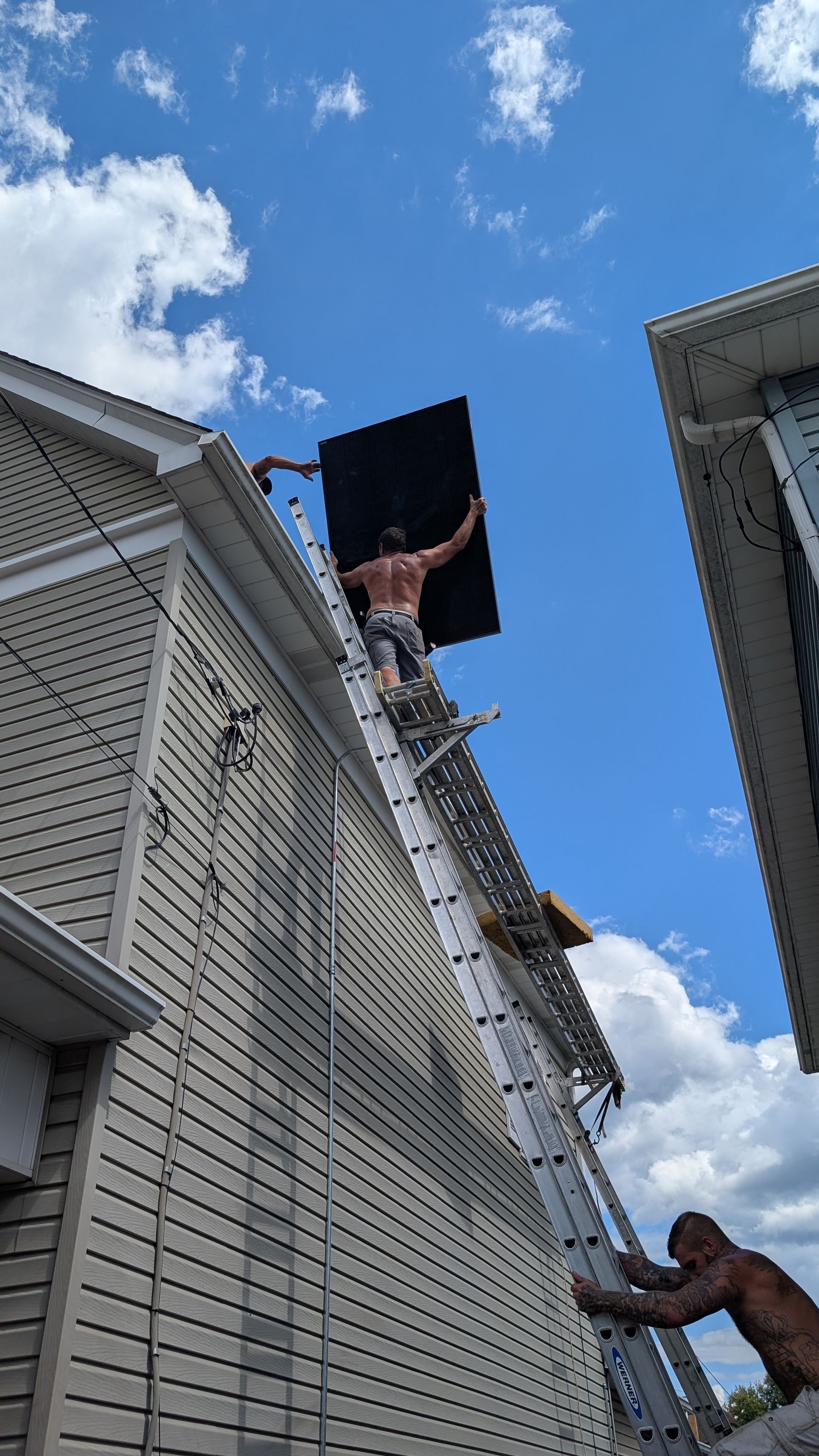 Two men on a ladder placing a black rectangle on a roof, blue sky with clouds.