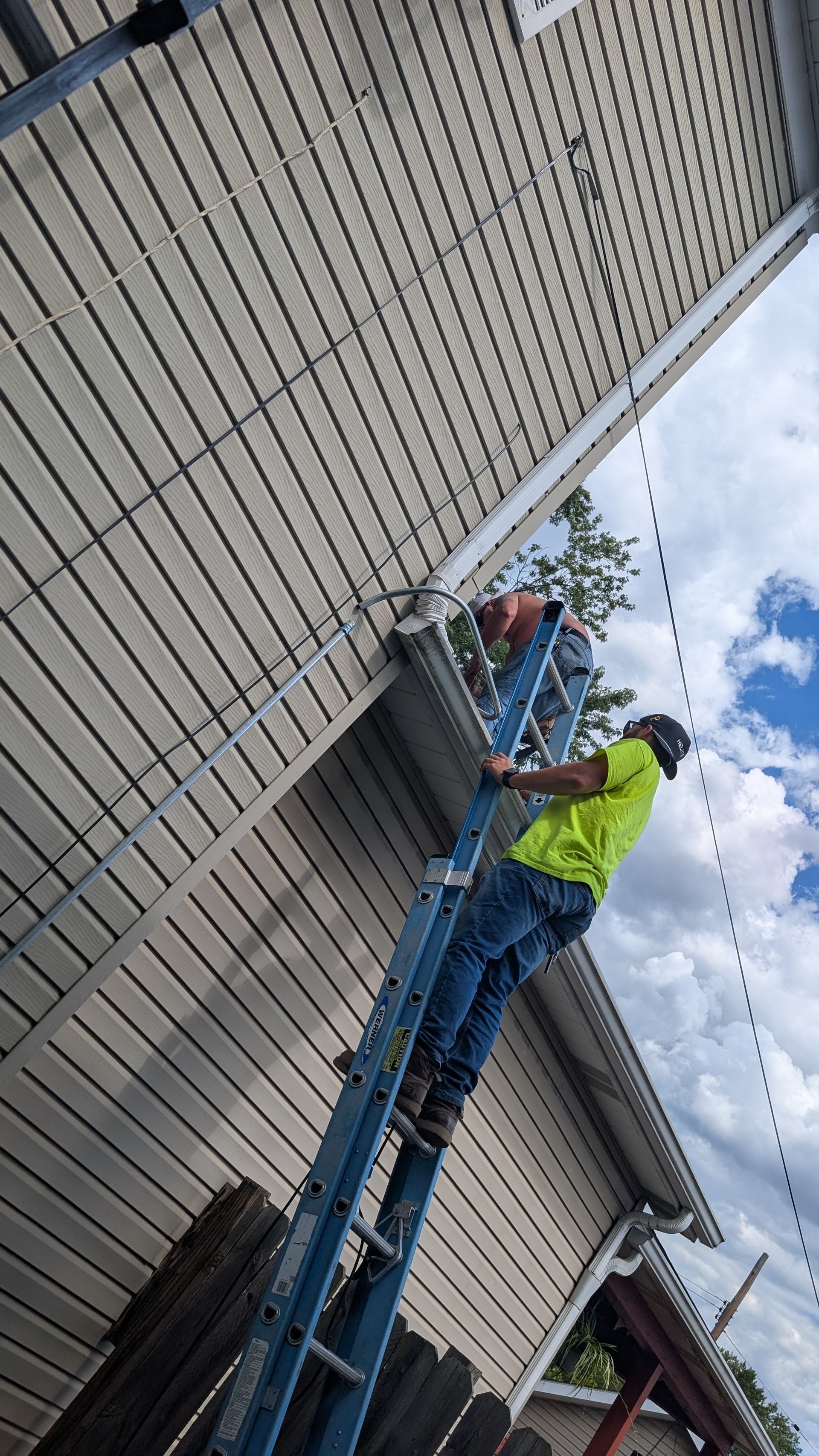 Man on ladder repairs house siding. Green shirt, blue jeans. Exterior setting, cloudy sky.