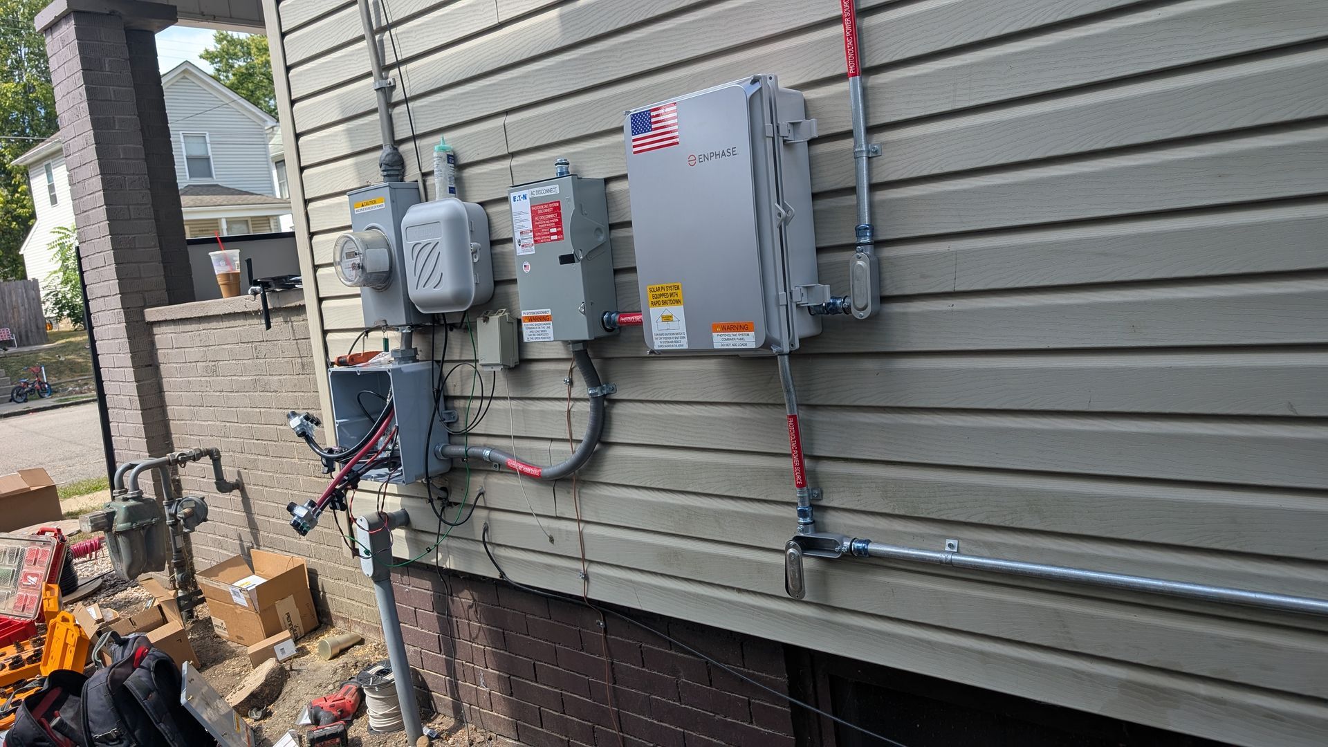Electrical equipment mounted on the side of a house with gray siding. Conduit and wires are visible.