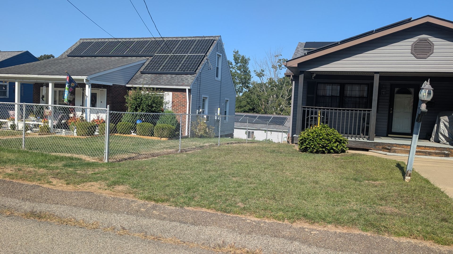 Two houses with solar panels on the roofs, separated by a chain-link fence and a grassy lawn, under a blue sky.