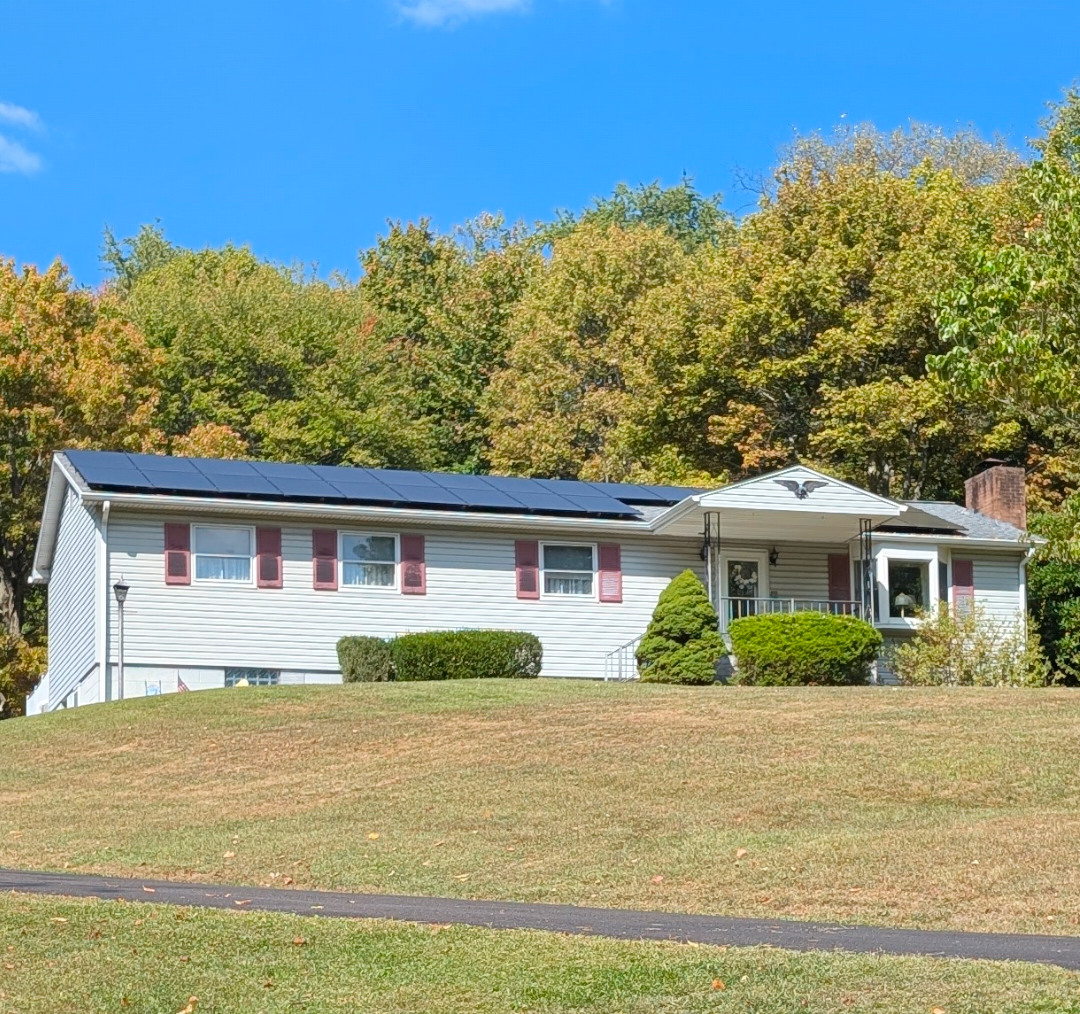 House on a grassy hill, blue sky above. White house, red shutters, trees in the background.