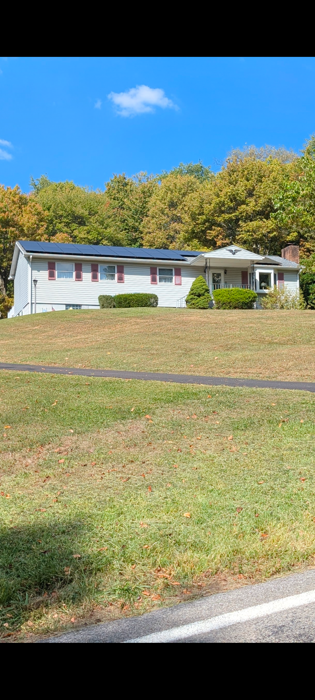 A white house with a blue sky in the background, solar panels on the roof, red shutters and a green yard.