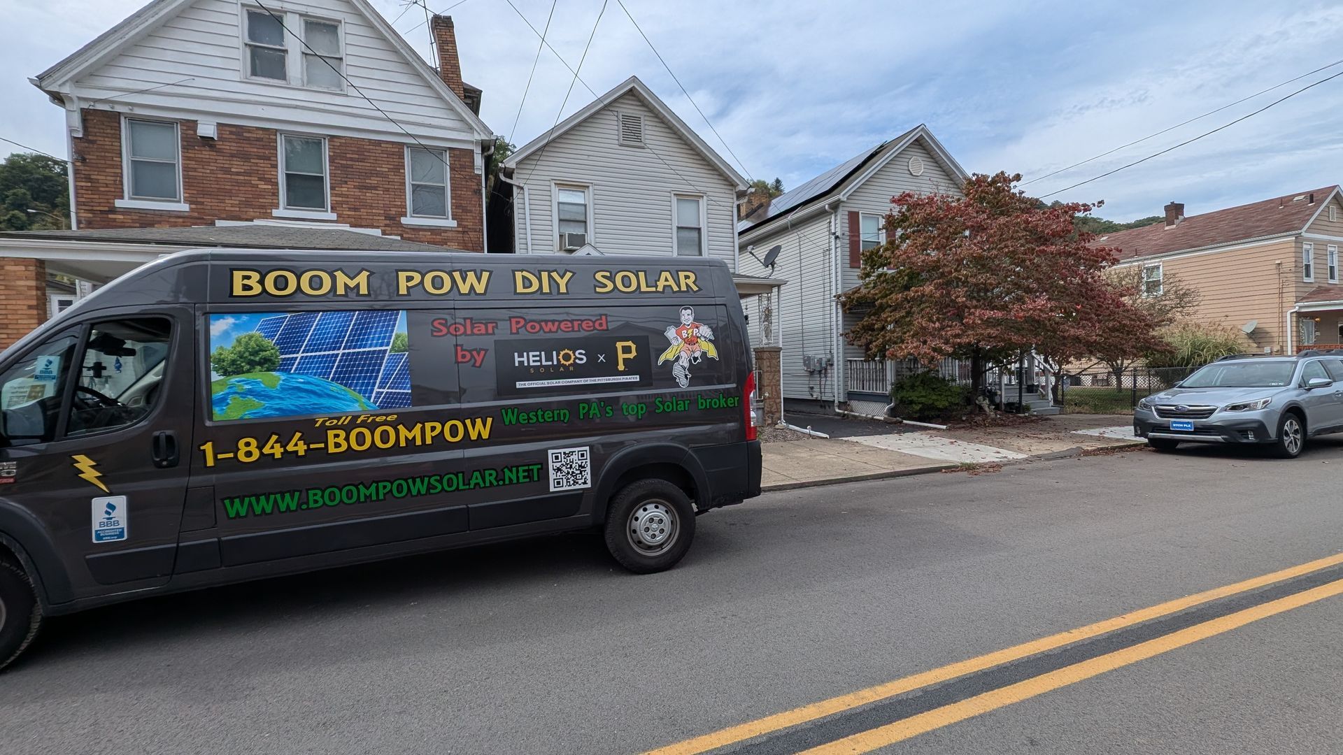 A dark van with solar panel graphics parked on a street in front of houses. The sky is overcast.