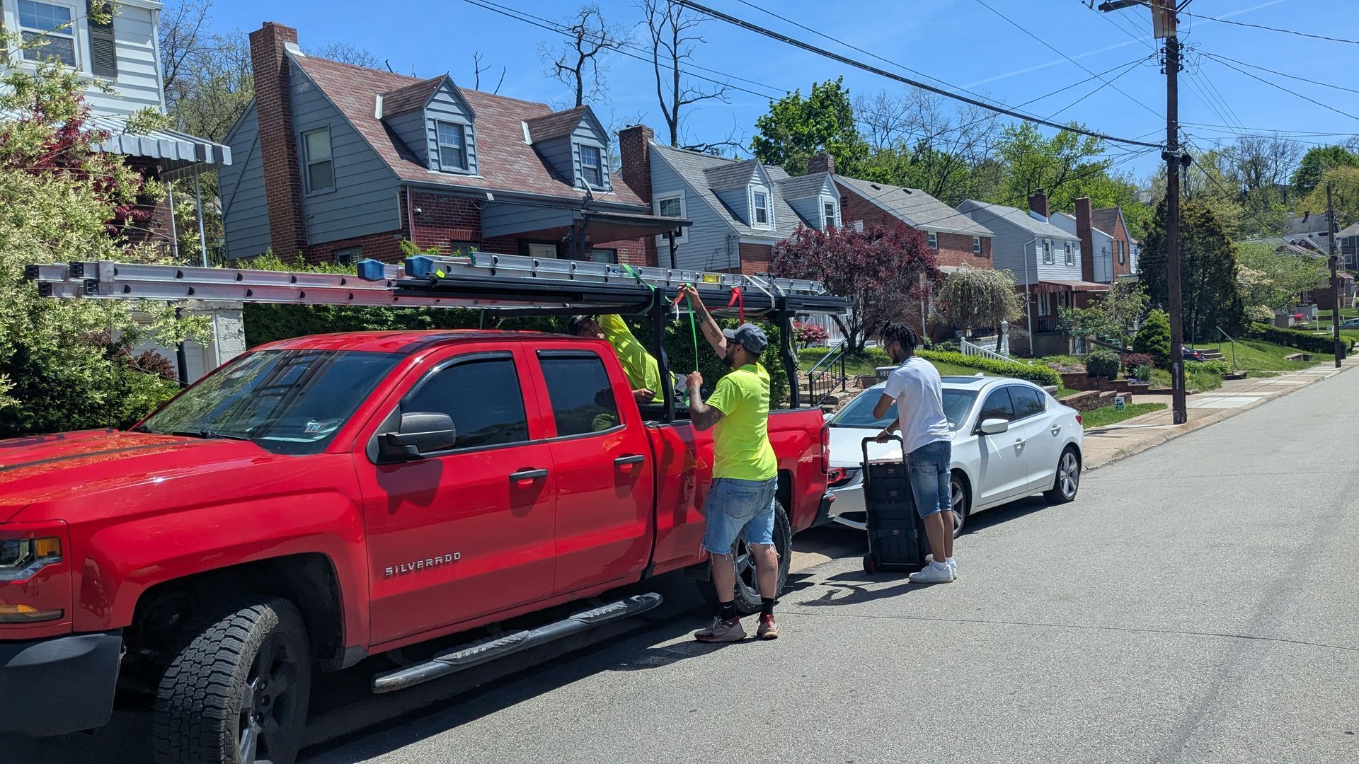 People loading a ladder onto a red truck parked on a street in front of houses on a sunny day. Two people standing next to a white car.