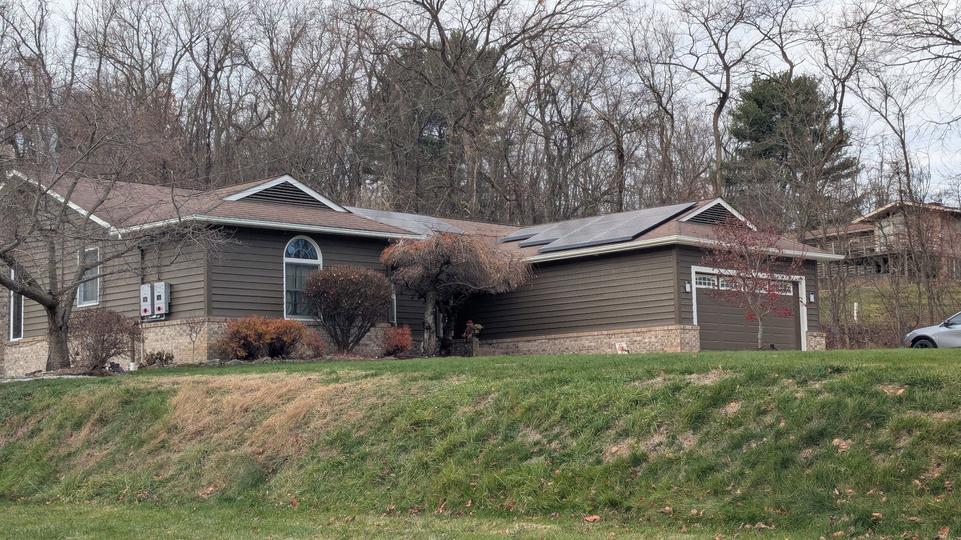 House with brown siding and roof, nestled among bare trees on a grassy hill.