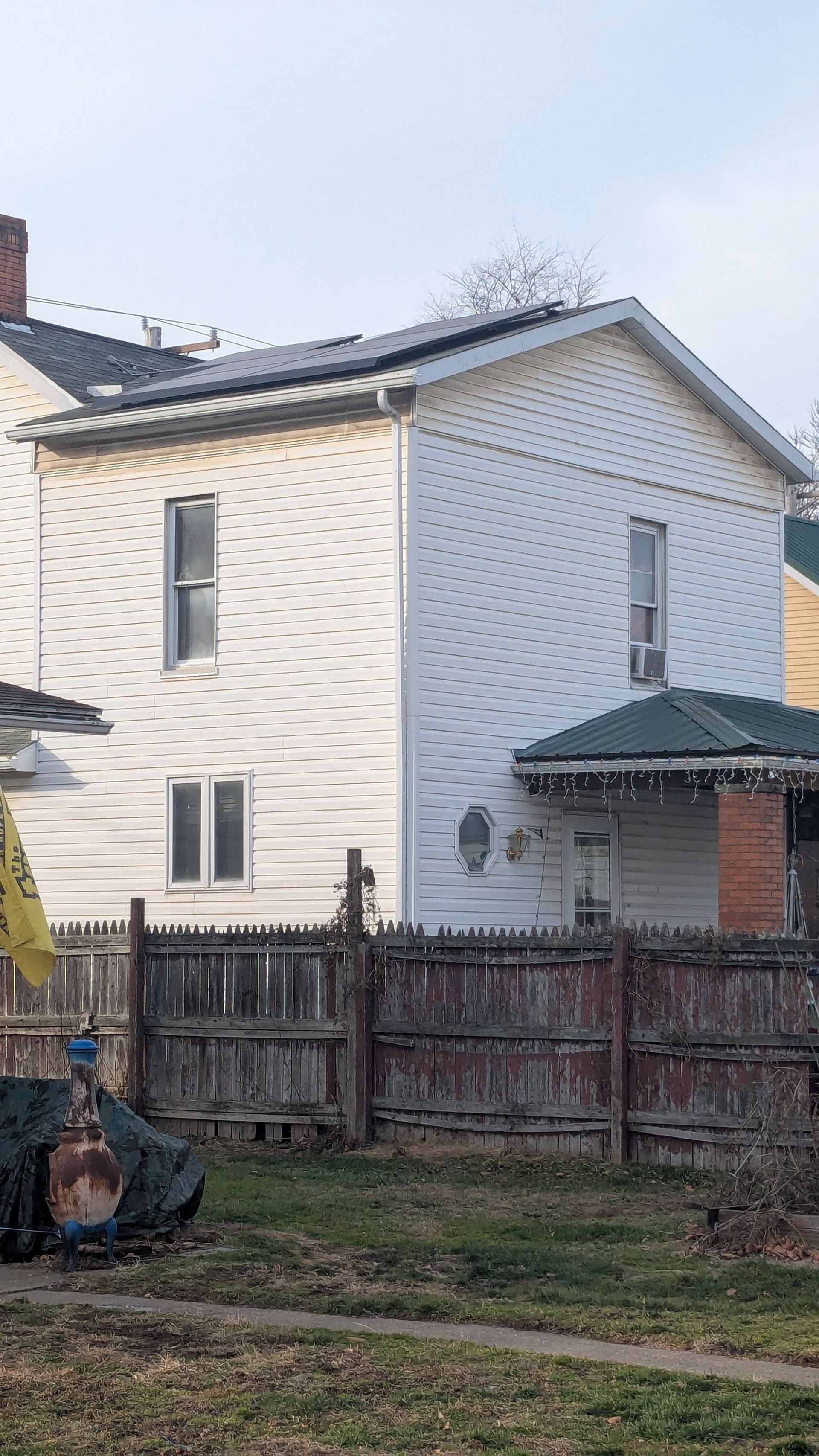 Two-story white house with solar panels on the roof, behind a weathered wooden fence.