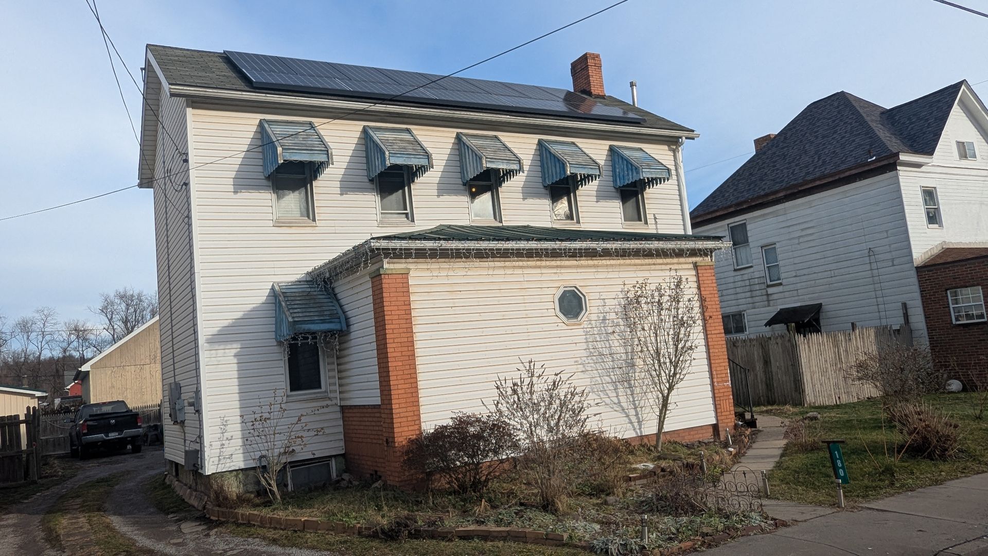 Two-story white house with solar panels, awnings, and a brick accent. Next to another house, both under a blue sky.