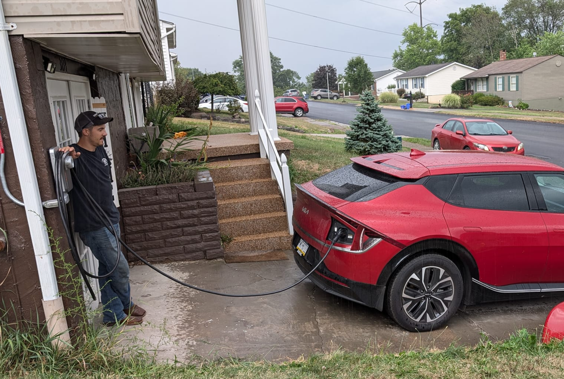 Man charging a red electric car outside a house. The charging cable extends from a wall-mounted charger to the car's port.