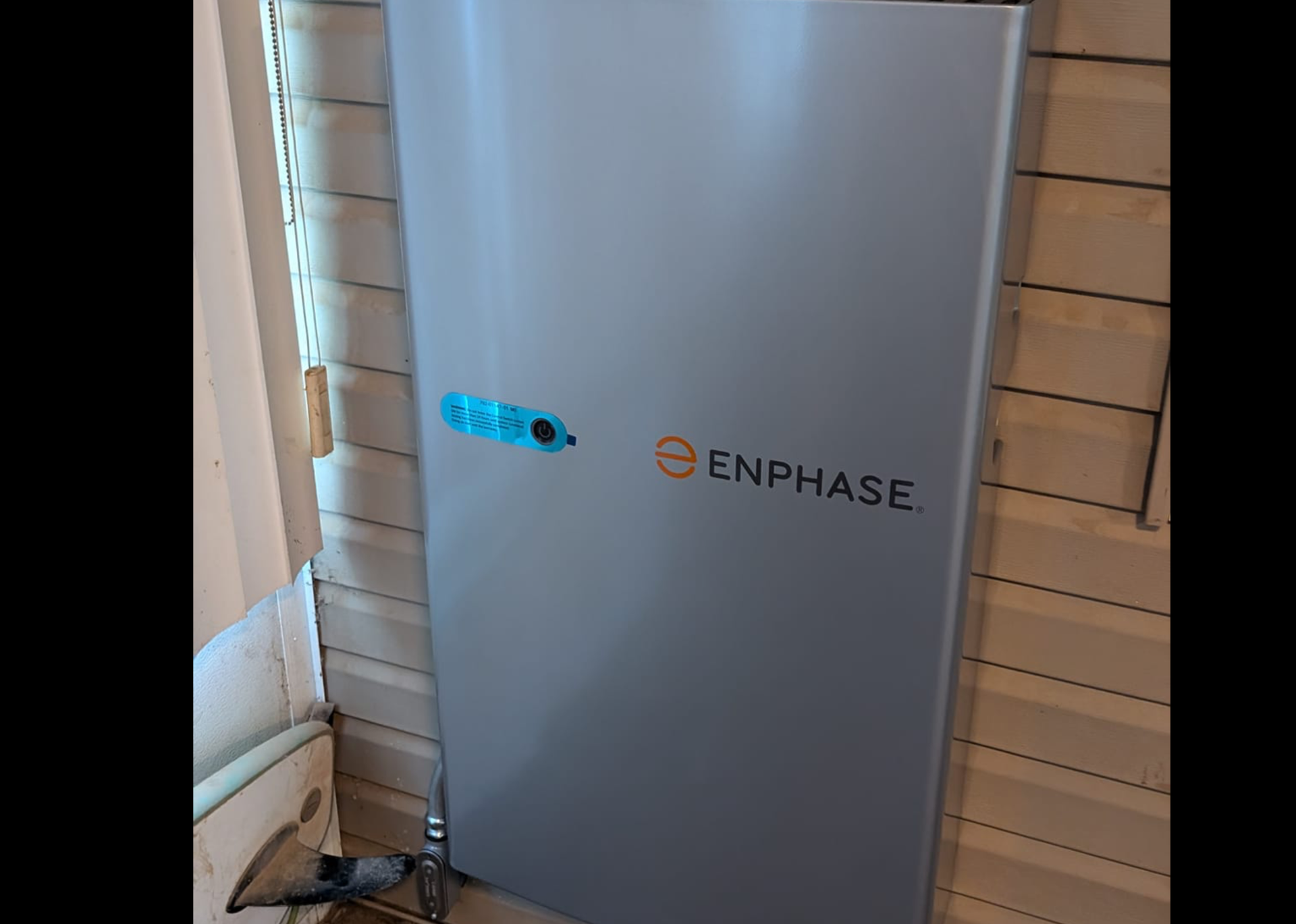 A gray Enphase energy storage system is mounted on a wall. A small blue oval is visible near the top.