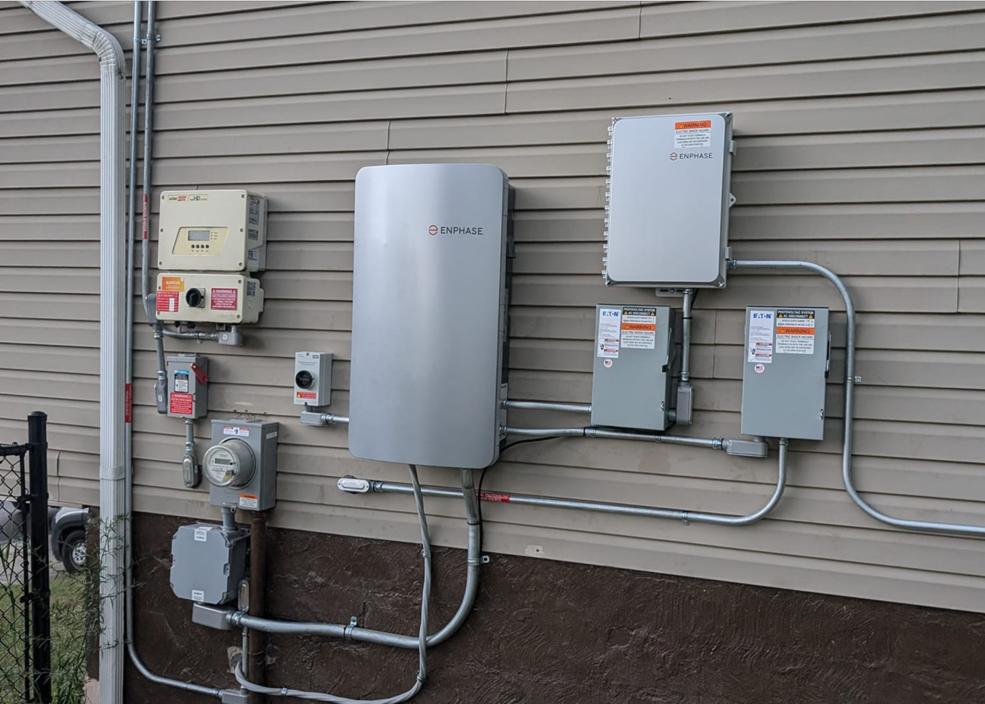 Exterior view of a house with solar panel equipment installed on the side. Various electrical boxes and wiring are attached to the light-colored siding.