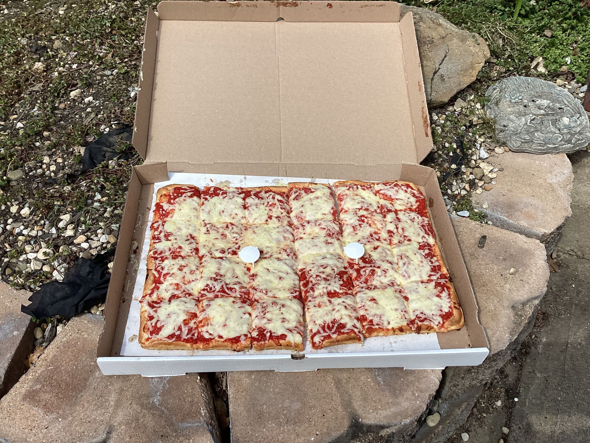 A square-cut cheese pizza in an open cardboard box, resting on a stone surface outdoors.