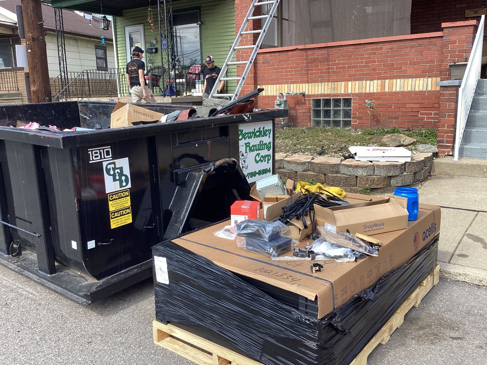 A black dumpster sits beside a pallet of building materials and a ladder in front of a house undergoing renovations.
