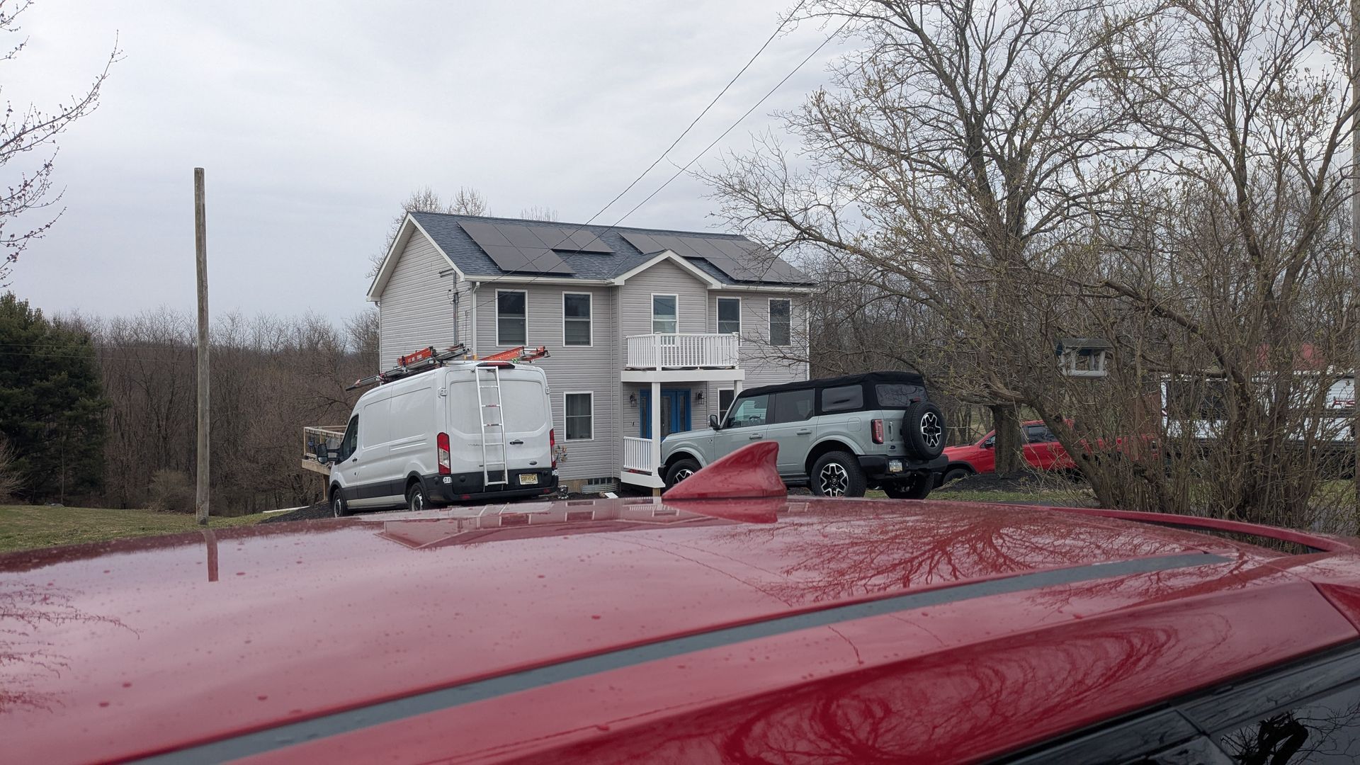 Red car parked in front of a two-story house. A white van and a grey SUV are also parked nearby. The sky is overcast.