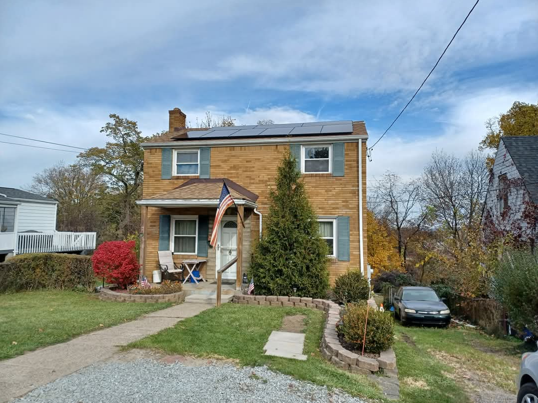 Two-story brick house with solar panels, blue shutters, small porch, and a car in the driveway on a sunny day.