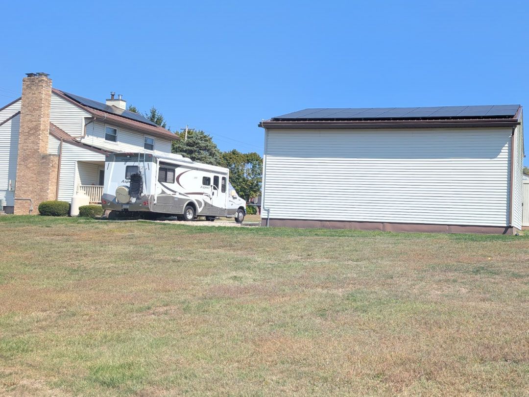 RV parked next to a house and a white shed on a grassy lawn under a blue sky.