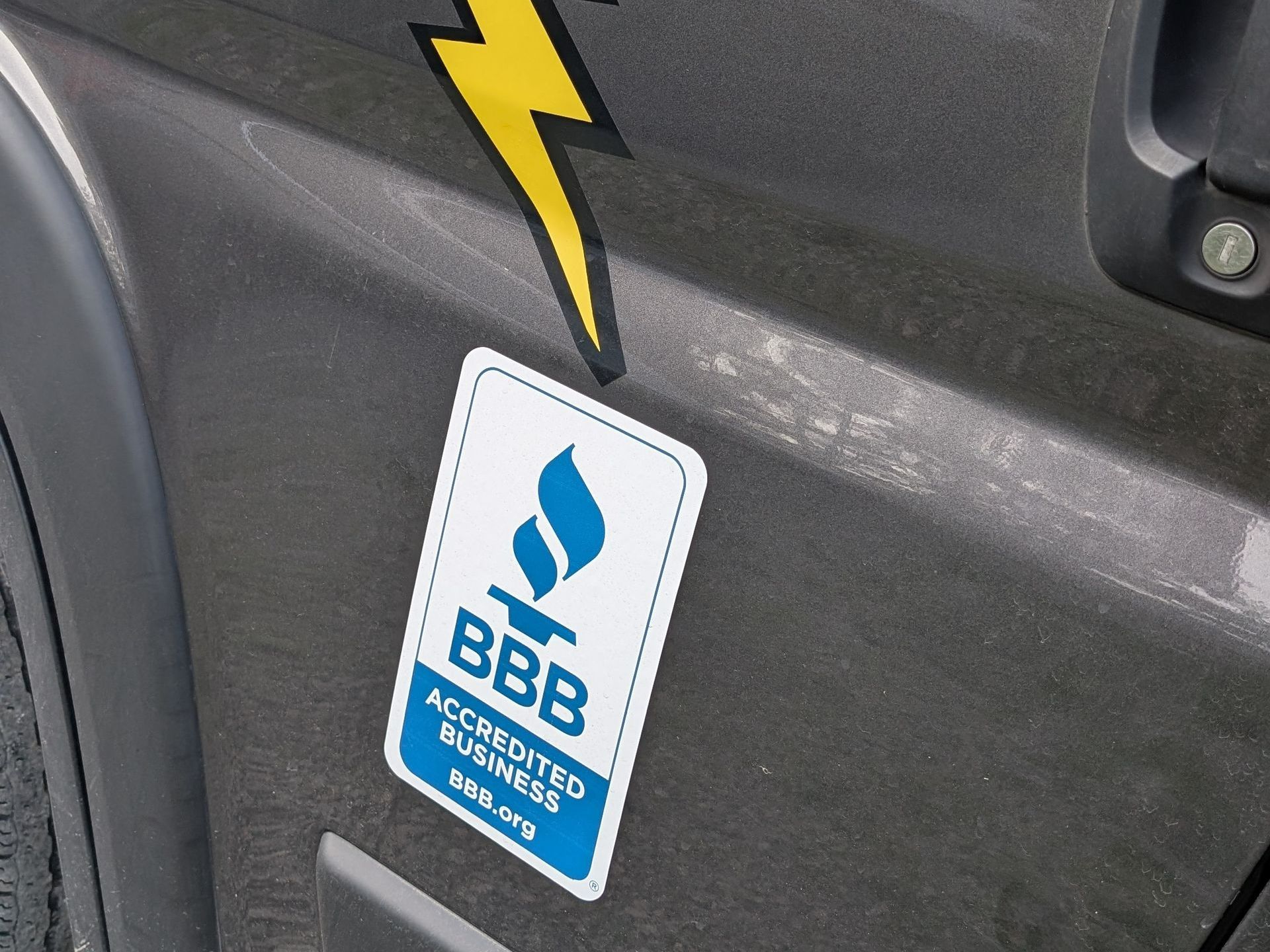 Close-up of a vehicle showing a blue and white Better Business Bureau (BBB) Accredited Business sticker next to a yellow lightning bolt graphic.