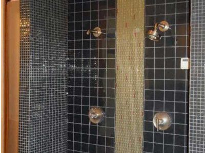 Black tiled shower with gold mosaic accent; multiple showerheads.