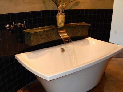 White bathtub with waterfall faucet, black tile backsplash, and decorative display.