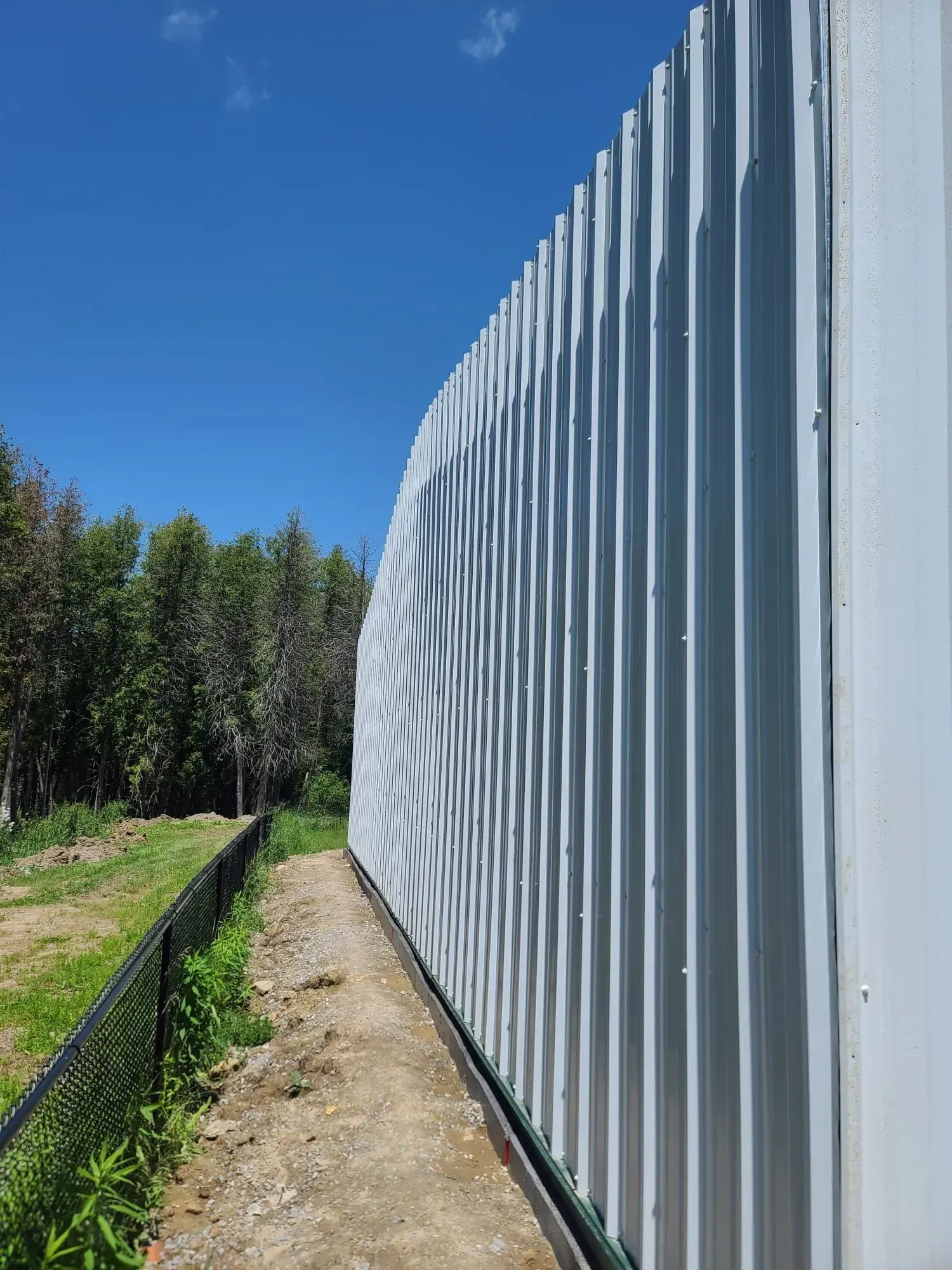 A large white metal fence is surrounded by trees and a dirt path.