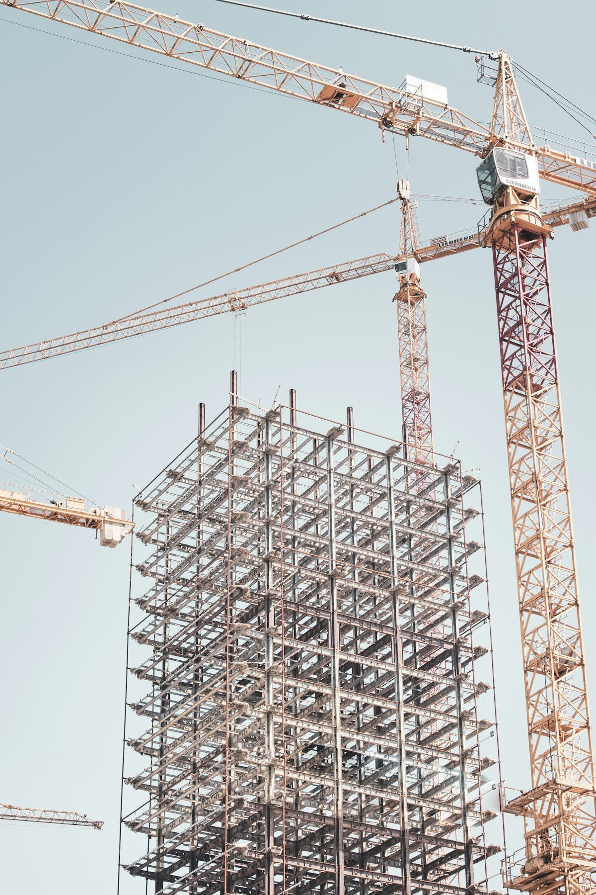 Two construction cranes beside a high-rise building frame under construction against a pale sky