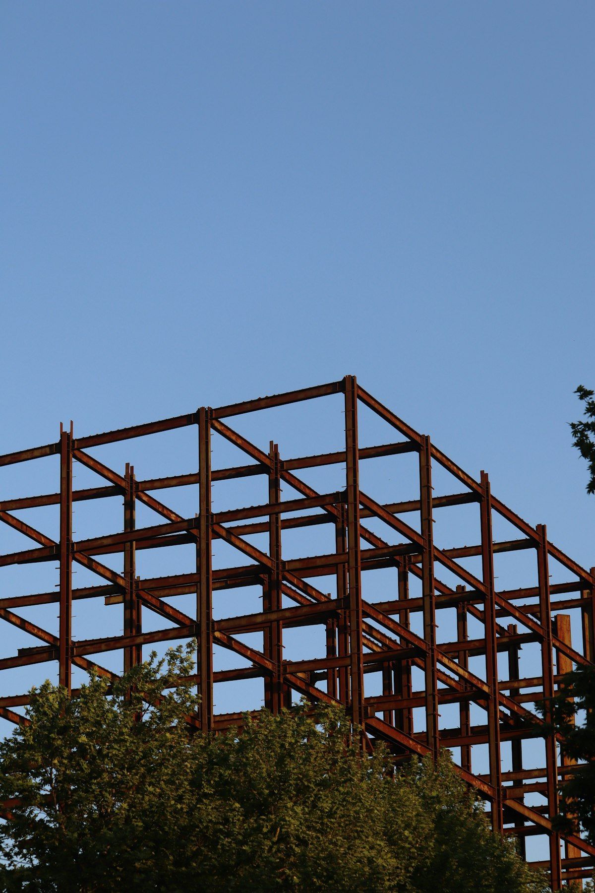 Rust-colored steel framework under construction against a clear blue sky, partly obscured by trees