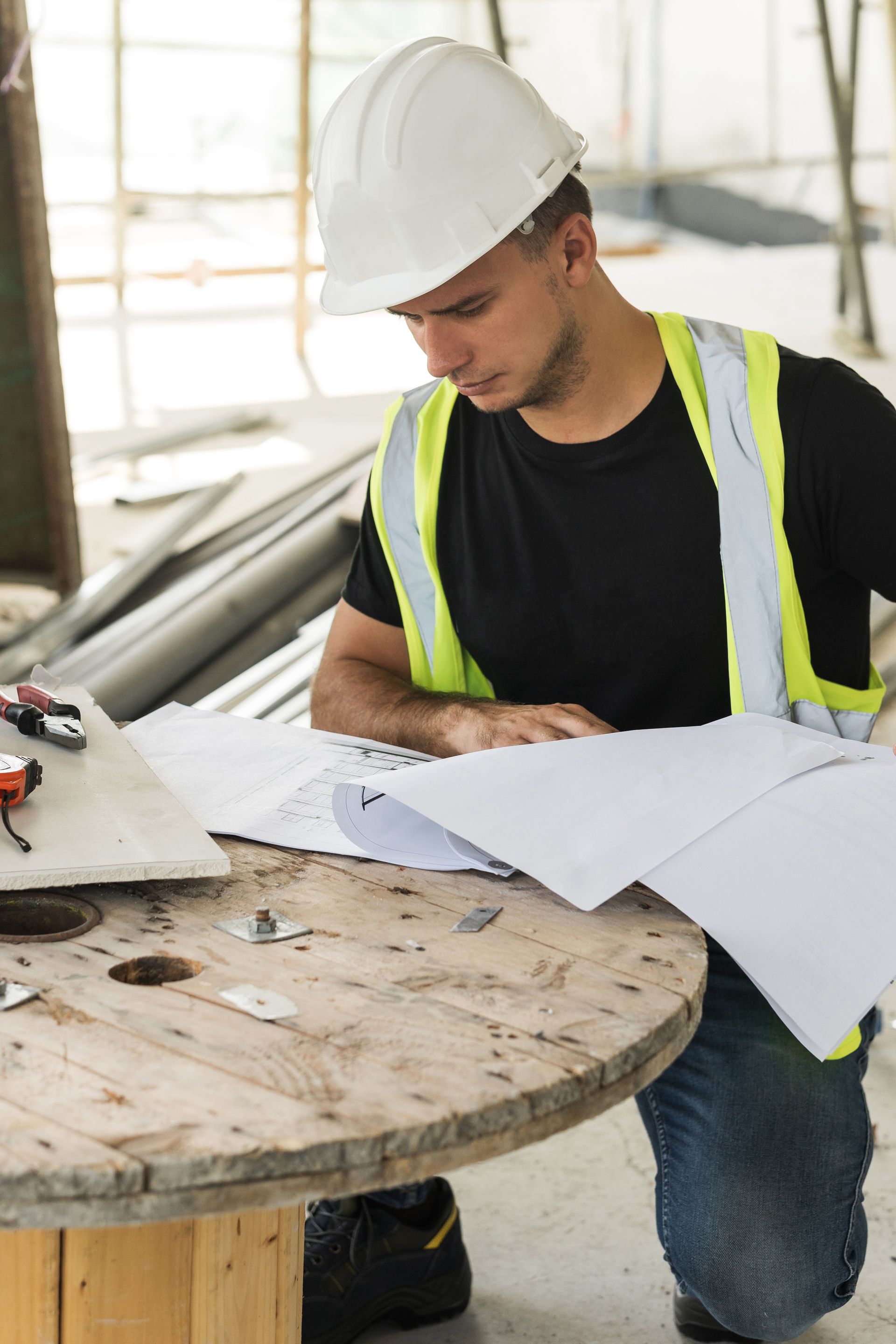 A construction worker is sitting on a wooden spool looking at a blueprint.