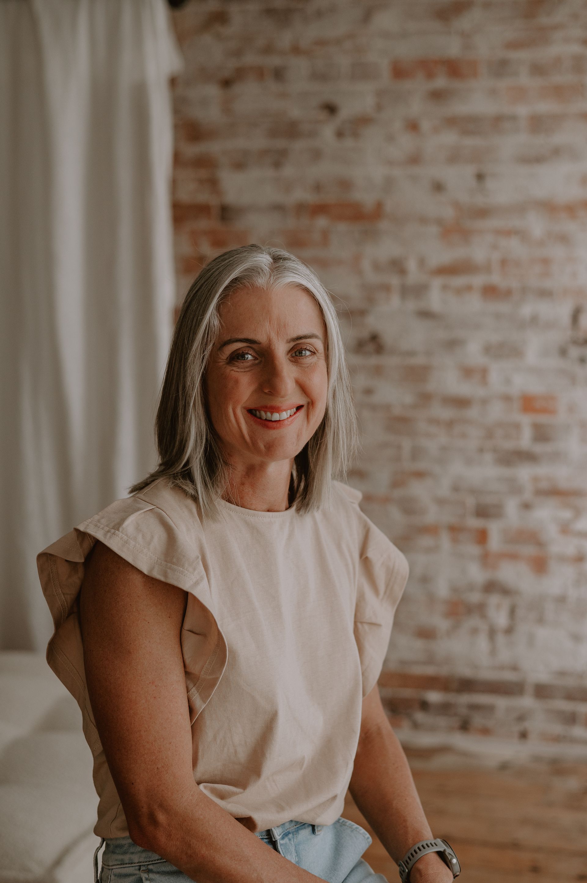 Woman with gray hair smiles, sitting against a brick wall; wearing a beige top and watch.