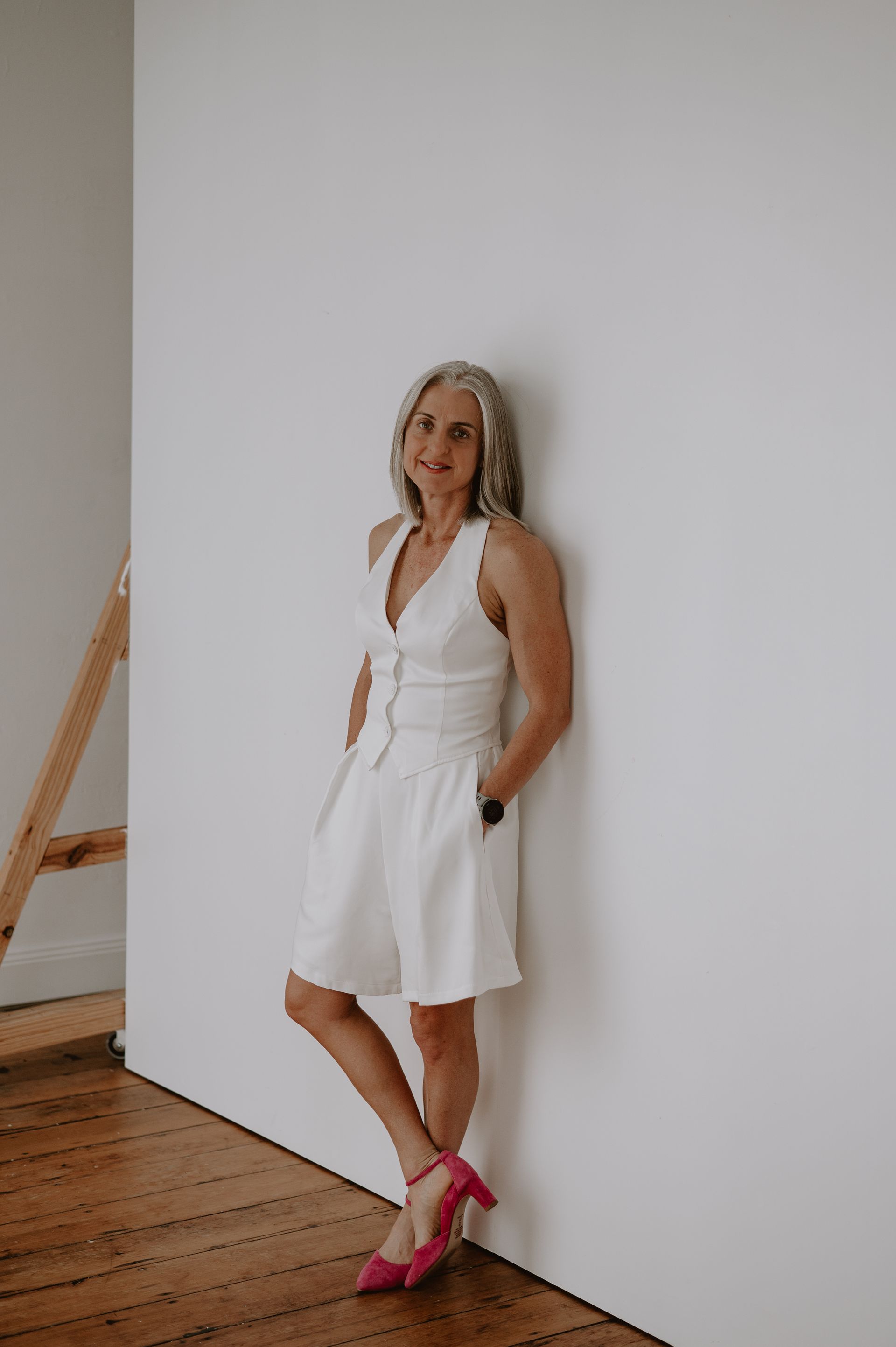 Woman with gray hair wearing white outfit and pink heels, leaning against a white wall. Woman with gray hair wearing white outfit and pink heels, leaning against a white wall.