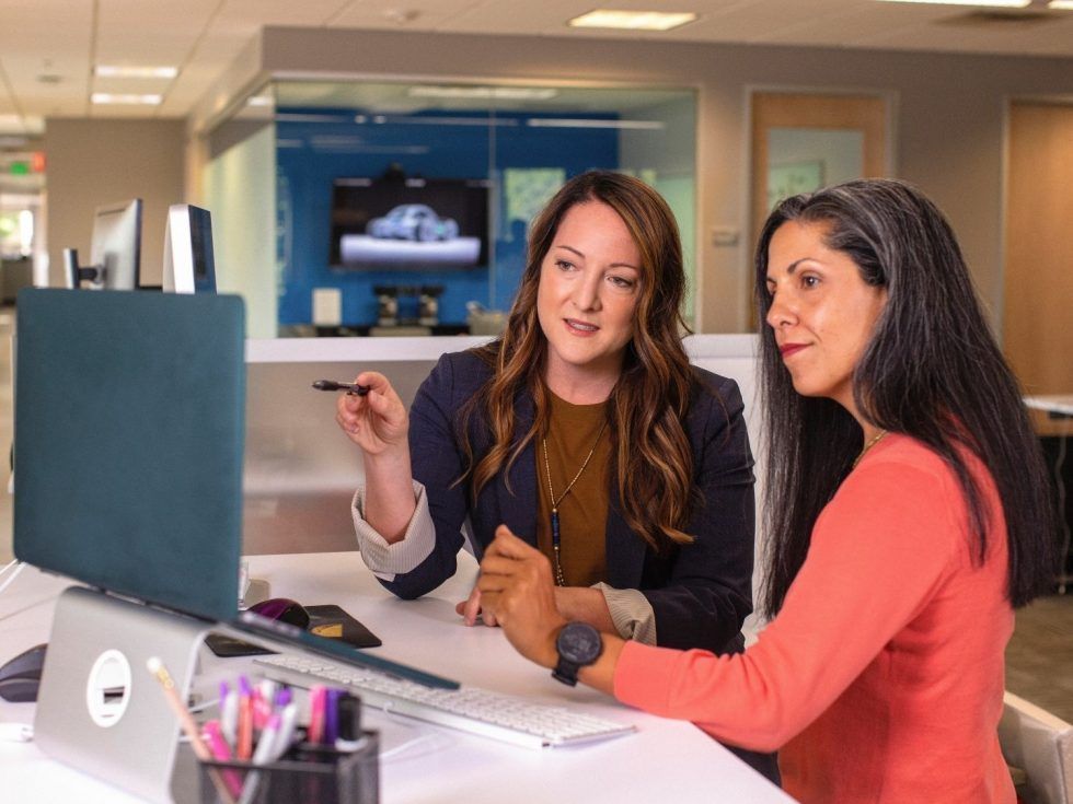 Two women are sitting at a desk looking at a laptop computer.