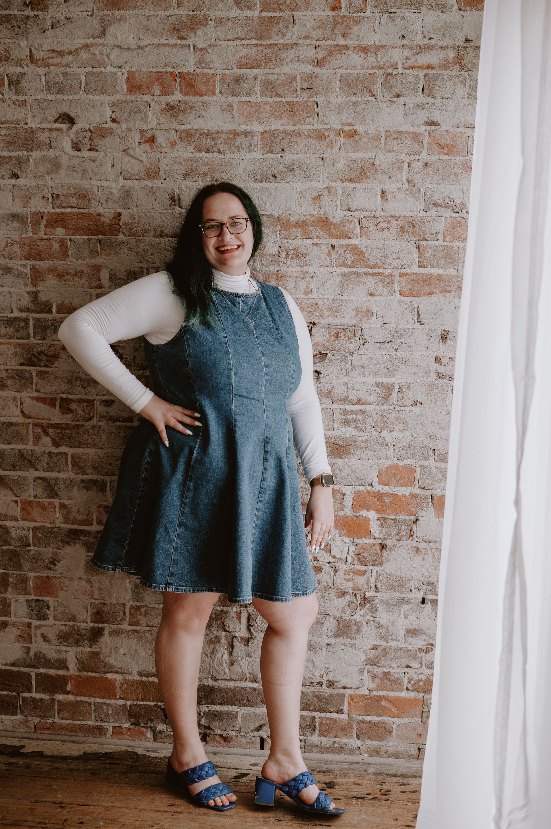 Woman in denim dress and turtleneck, posing against a brick wall, wearing glasses and blue sandals.
