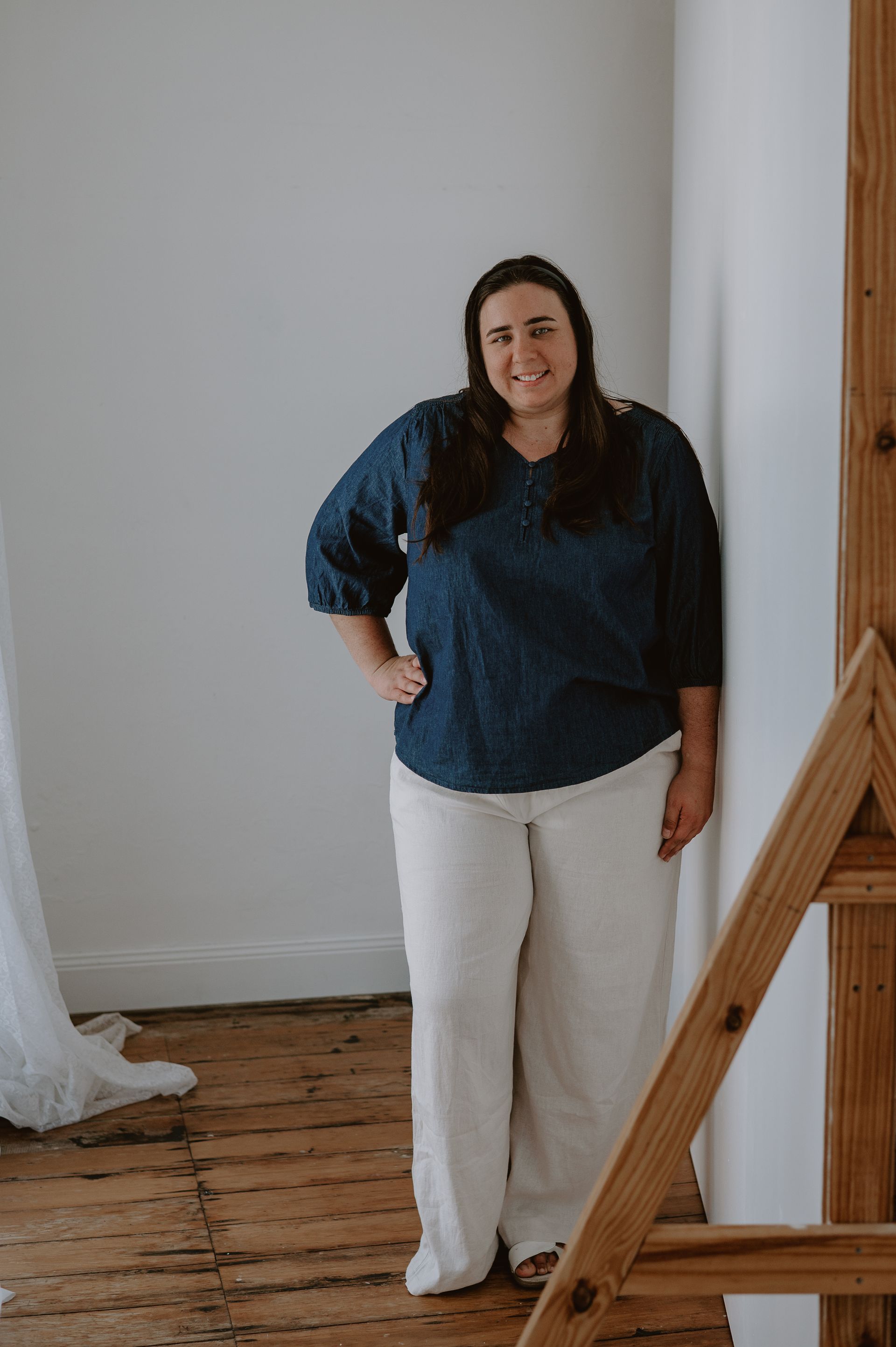 Woman in denim top and white pants leans against a white wall, smiling.