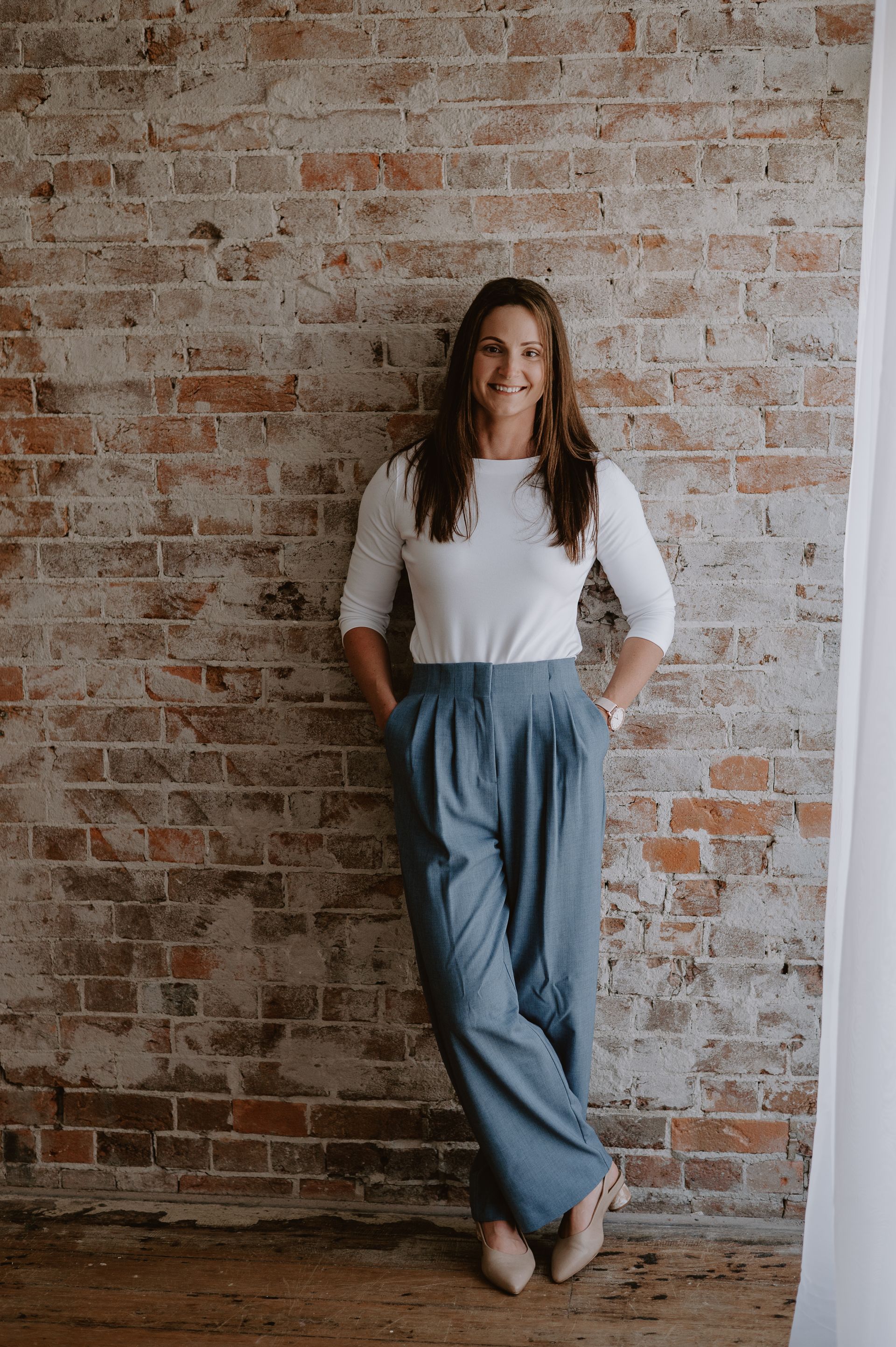 Woman in white shirt and blue pants, leaning against a brick wall, smiling.