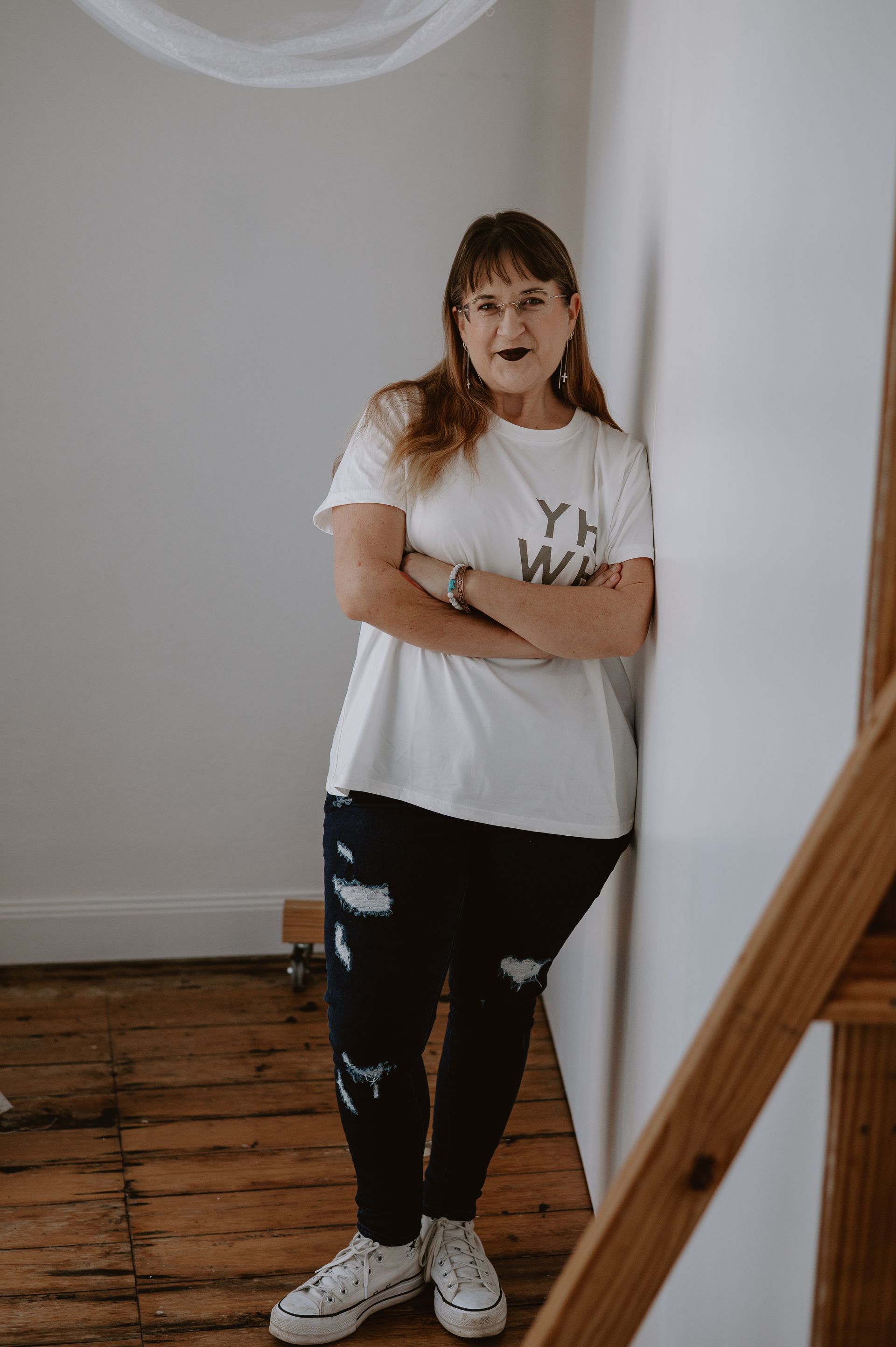 Woman with arms crossed, leaning against white wall, wearing white t-shirt and ripped jeans, converse shoes.