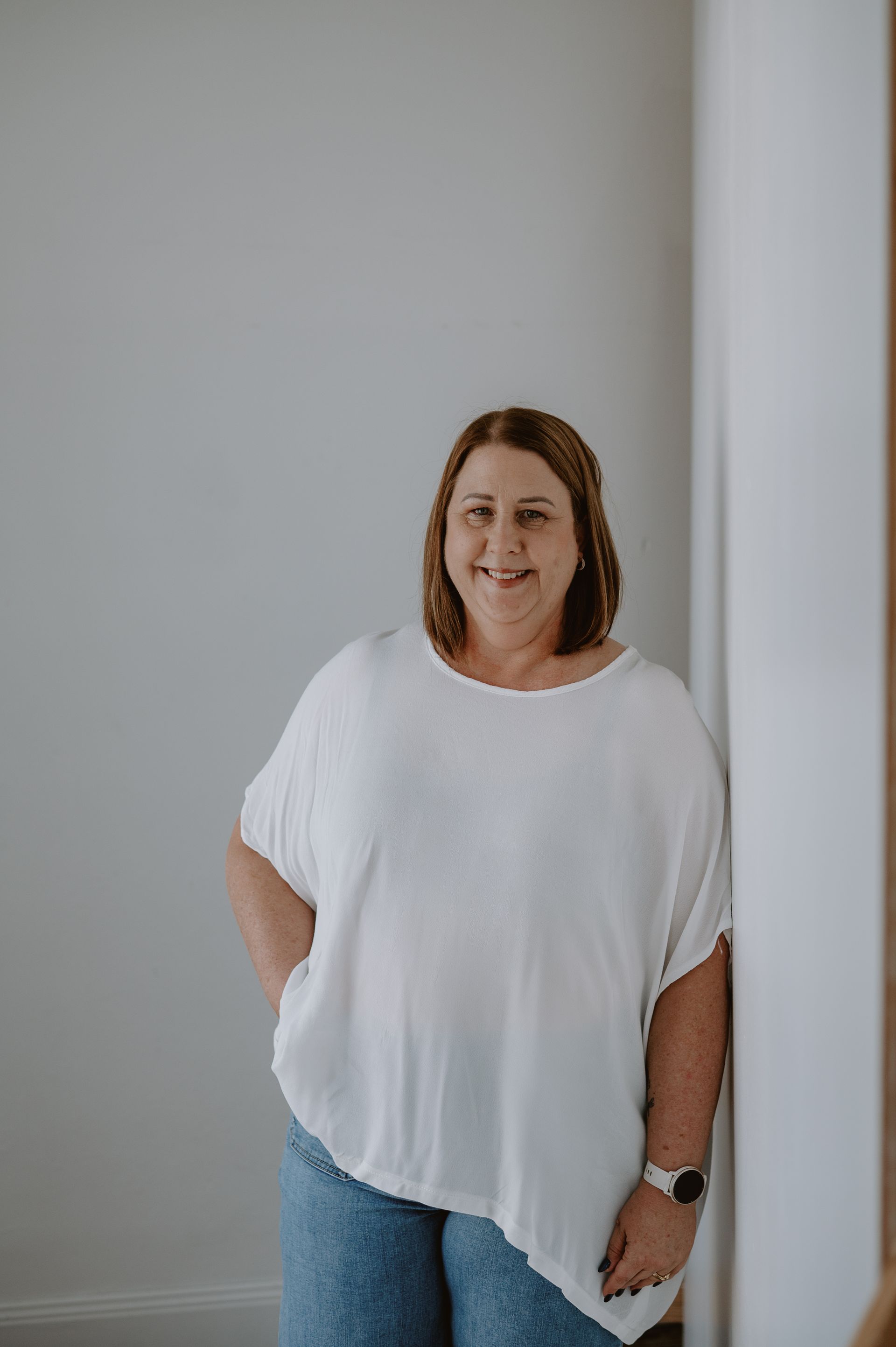 Woman in white top and jeans, smiling, leaning against a white wall.