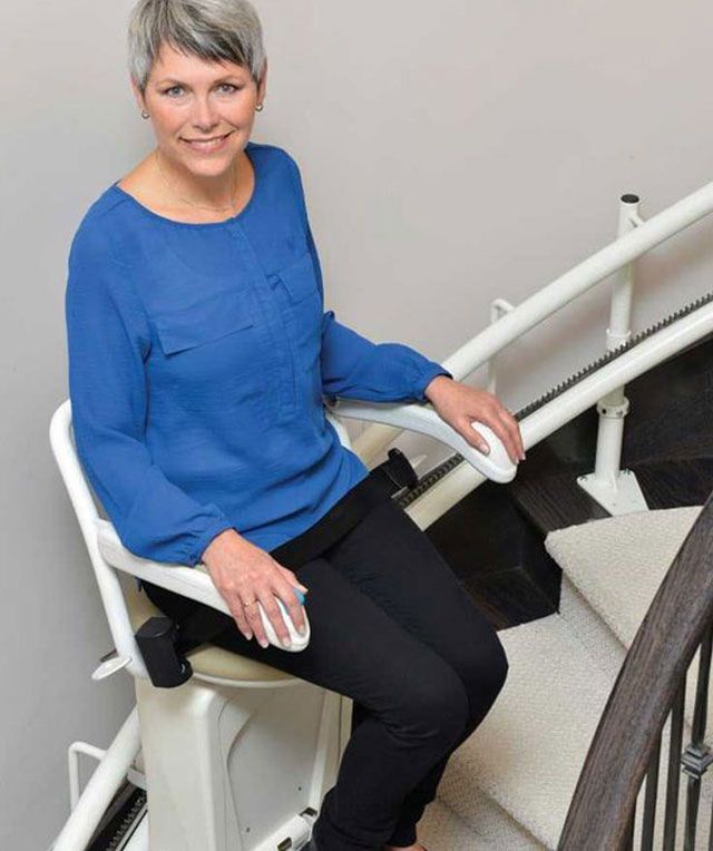 A Woman in A Blue Shirt Is Sitting on A Stair Lift — Master Lifts Cairns in Cairns, QLD