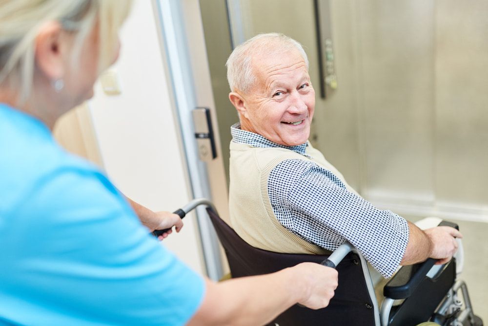 Happy Senior Man In Wheelchair Being Cared For By A Caregiver In Rehab — Master Lifts Cairns in Port Douglas, QLD